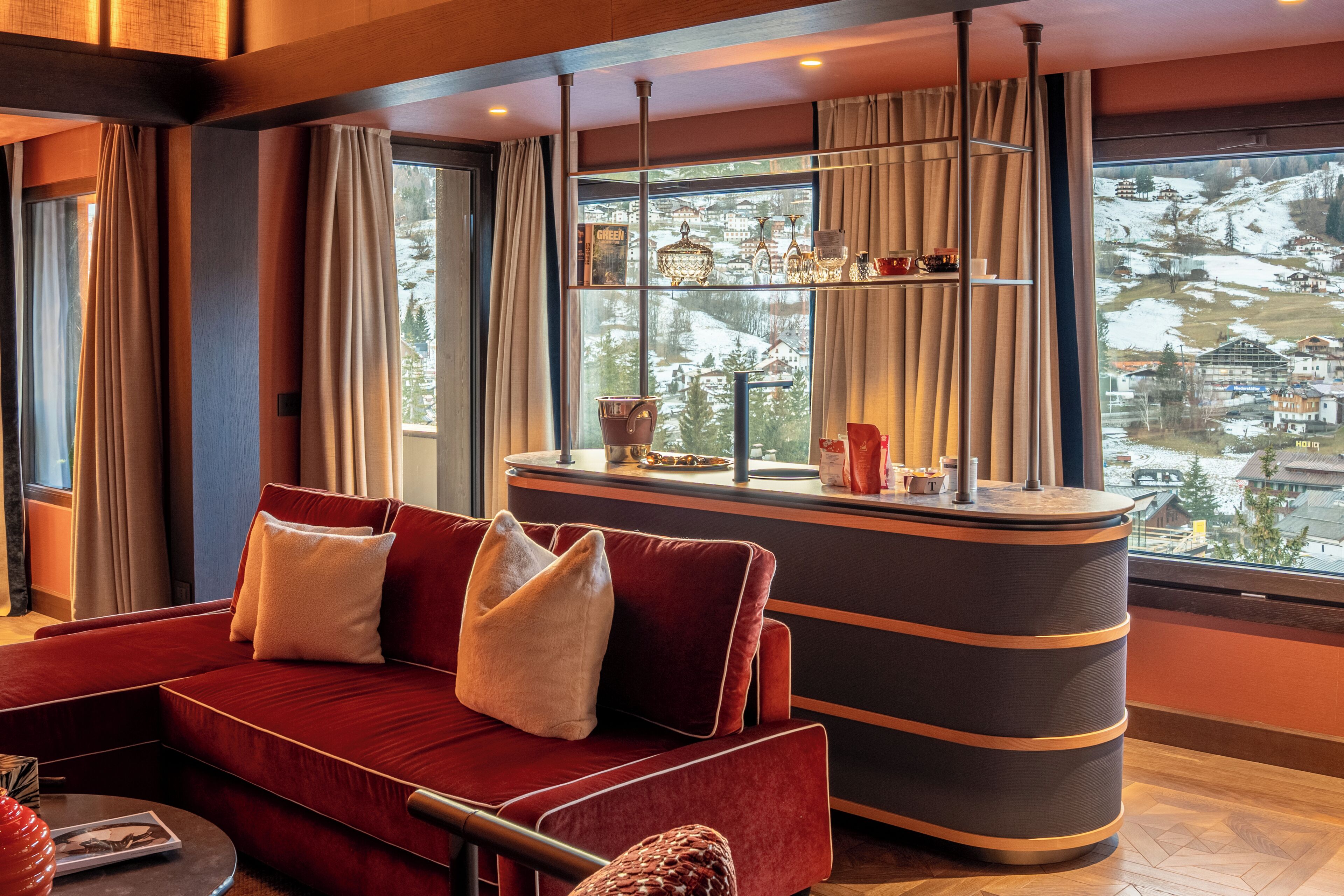 Une chambre d’hôtel moderne avec un canapé rouge, un bar au comptoir incurvé et une grande fenêtre donnant sur un paysage enneigé.