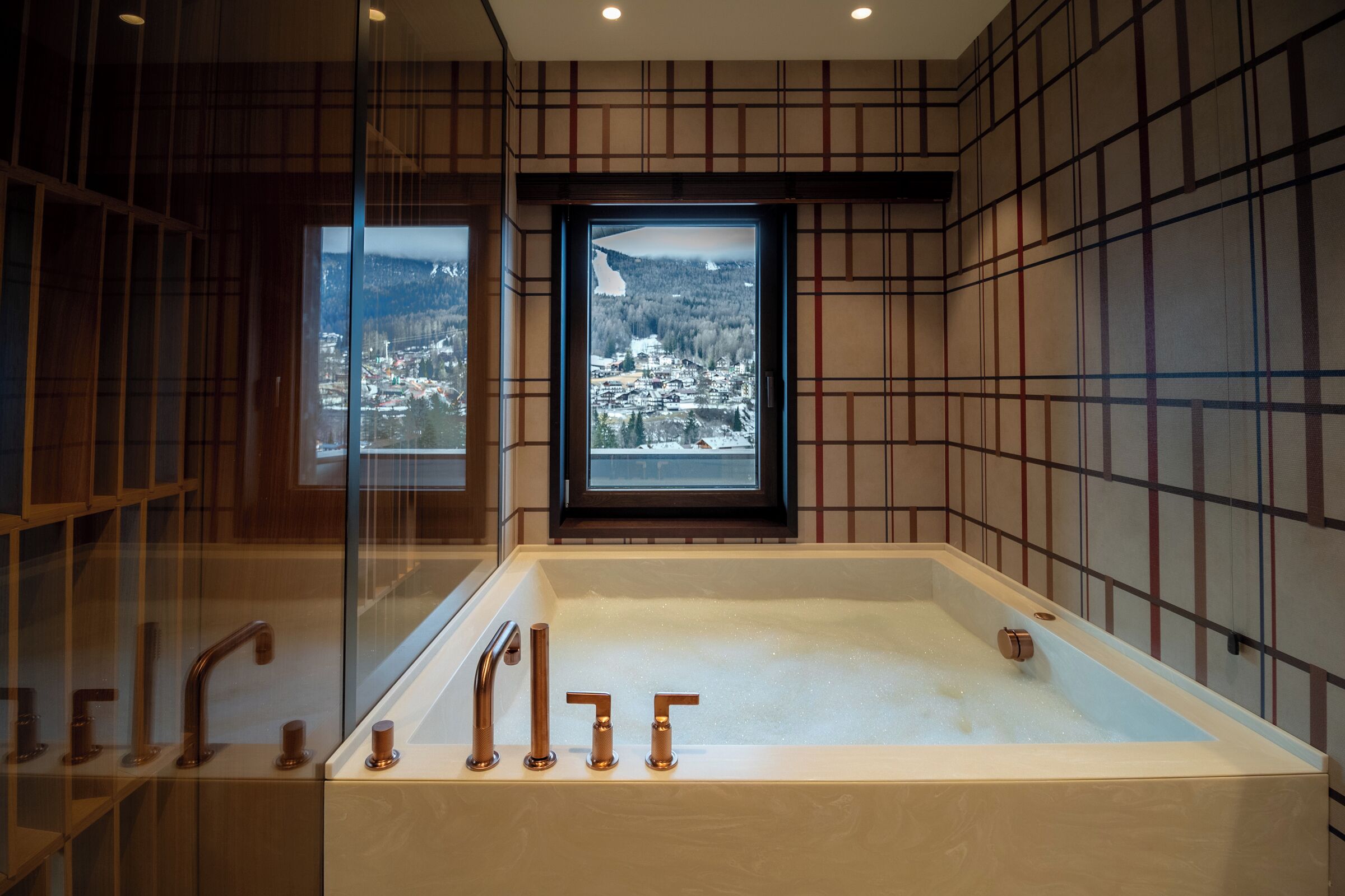 Une salle de bain moderne avec une grande baignoire, deux fenêtres donnant sur un paysage enneigé et un motif à carreaux sur un des murs.