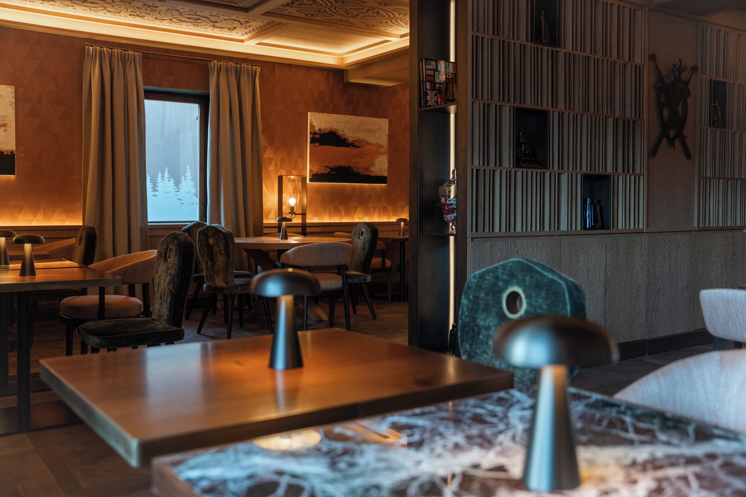 Una camera d'albergo con pavimento in legno, un'ampia finestra con tende, una scrivania con lampada, una sedia e una libreria con oggetti decorativi.