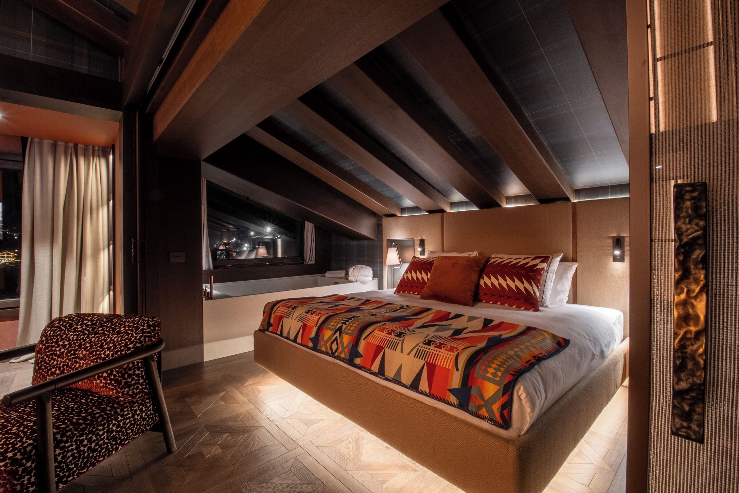 A modern bedroom with a large bed, a chair, and a view of the city at night.
