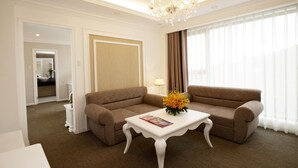 Ein Hotelzimmer mit einem braunen Ecksofa, einem weißen Couchtisch mit einer Zeitschrift, einem Kronleuchter und einem großen Fenster mit durchsichtigen Vorhängen.