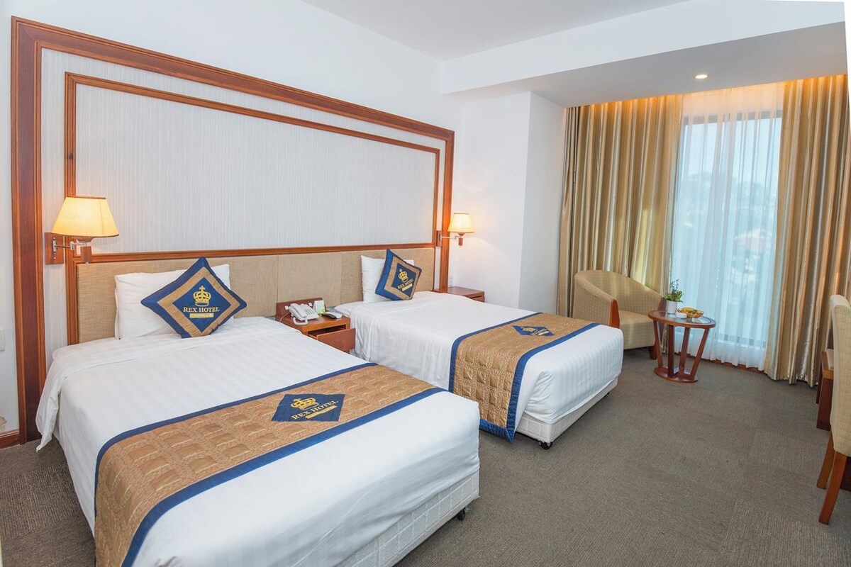 A hotel room with two beds, each with a headboard, bedside lamps, and a decorative pillow. There is a small table with a lamp and a plant, and a window with curtains.