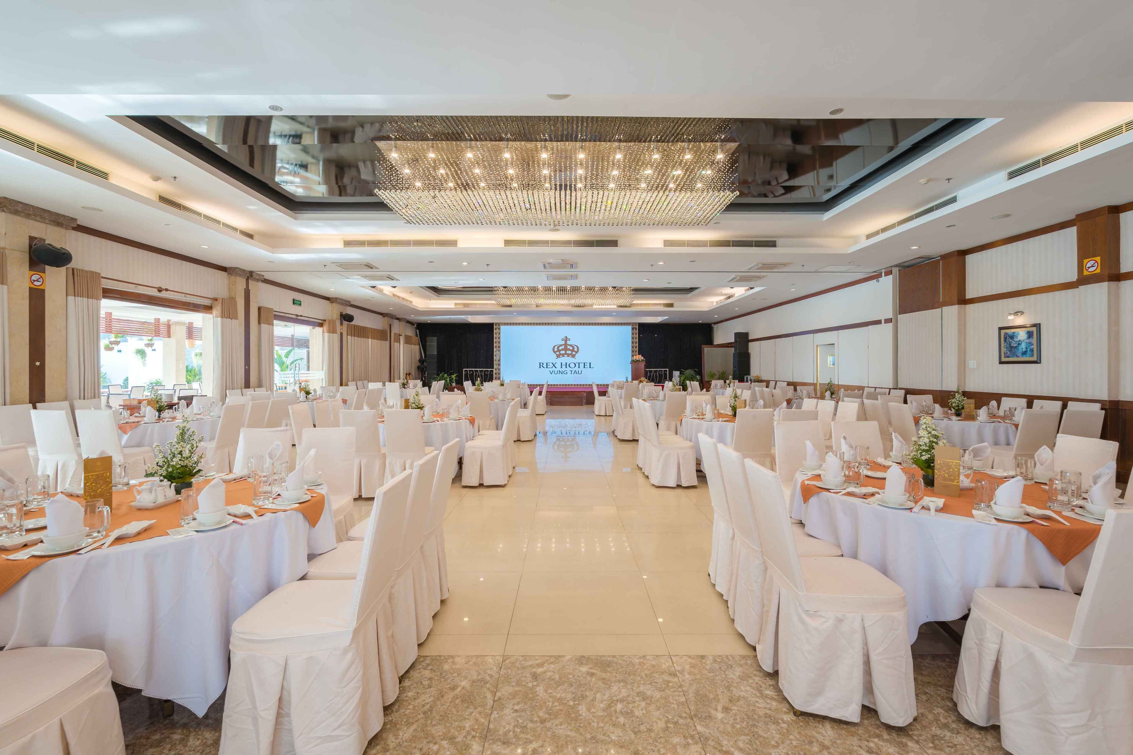 Un salón de banquetes con mesas redondas, sillas blancas y un escenario central con una pantalla de proyección.