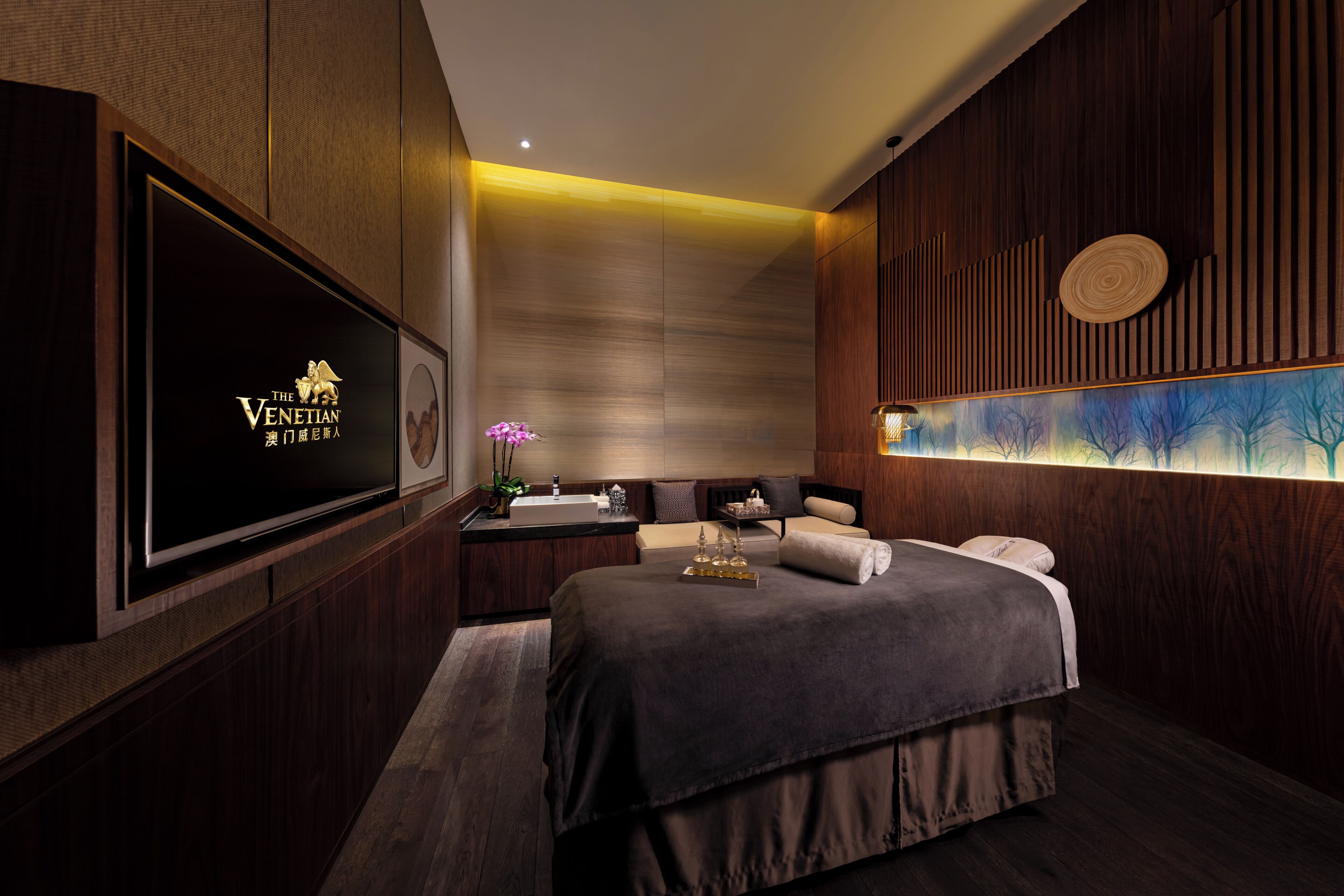 A modern hotel room with a large flat-screen TV, a bed with a dark-colored bedspread, a small bedside table, and a wall-mounted artwork.