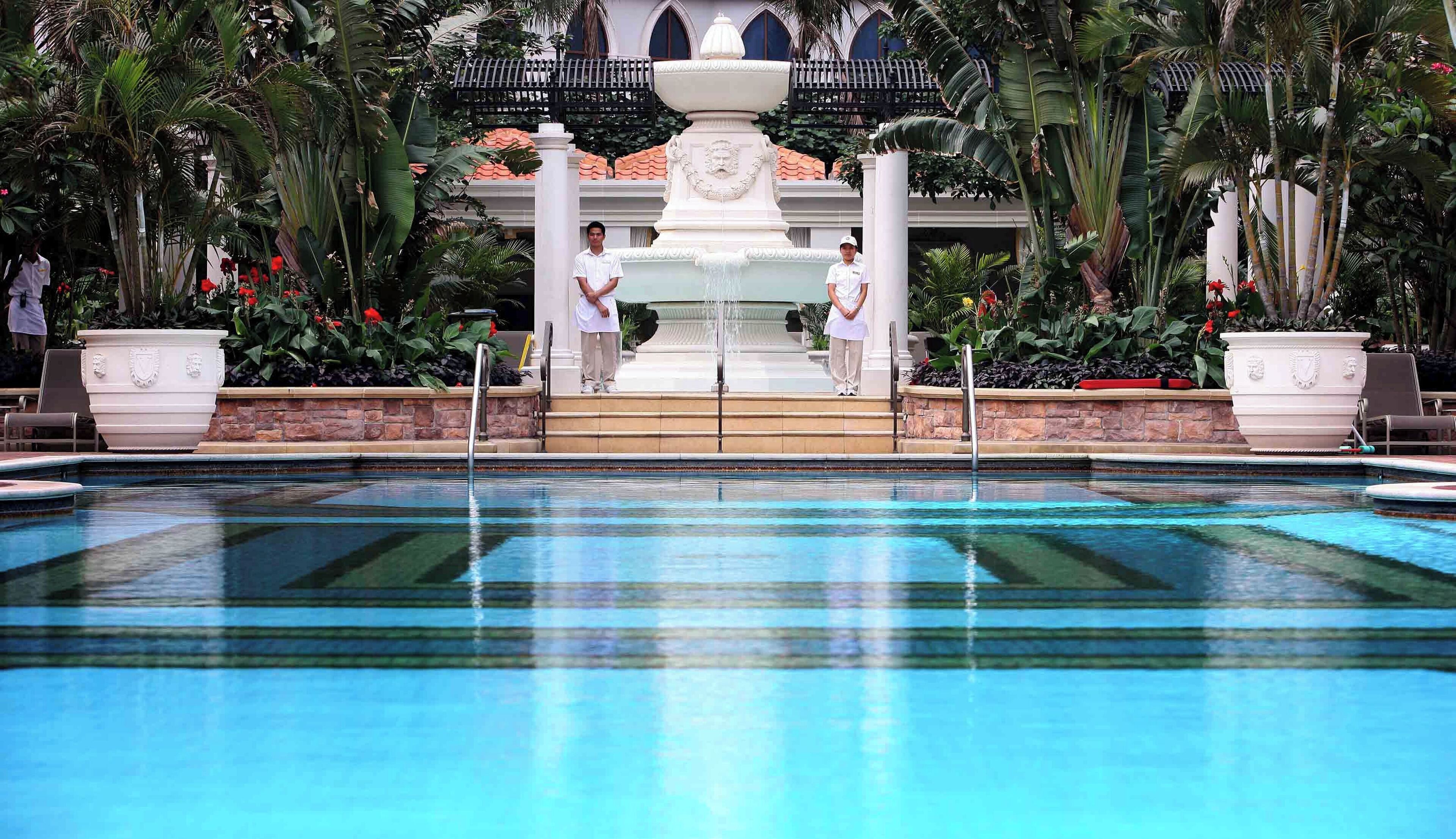 A pool area with a central fountain, surrounded by lush tropical plants and a white building with a balcony.