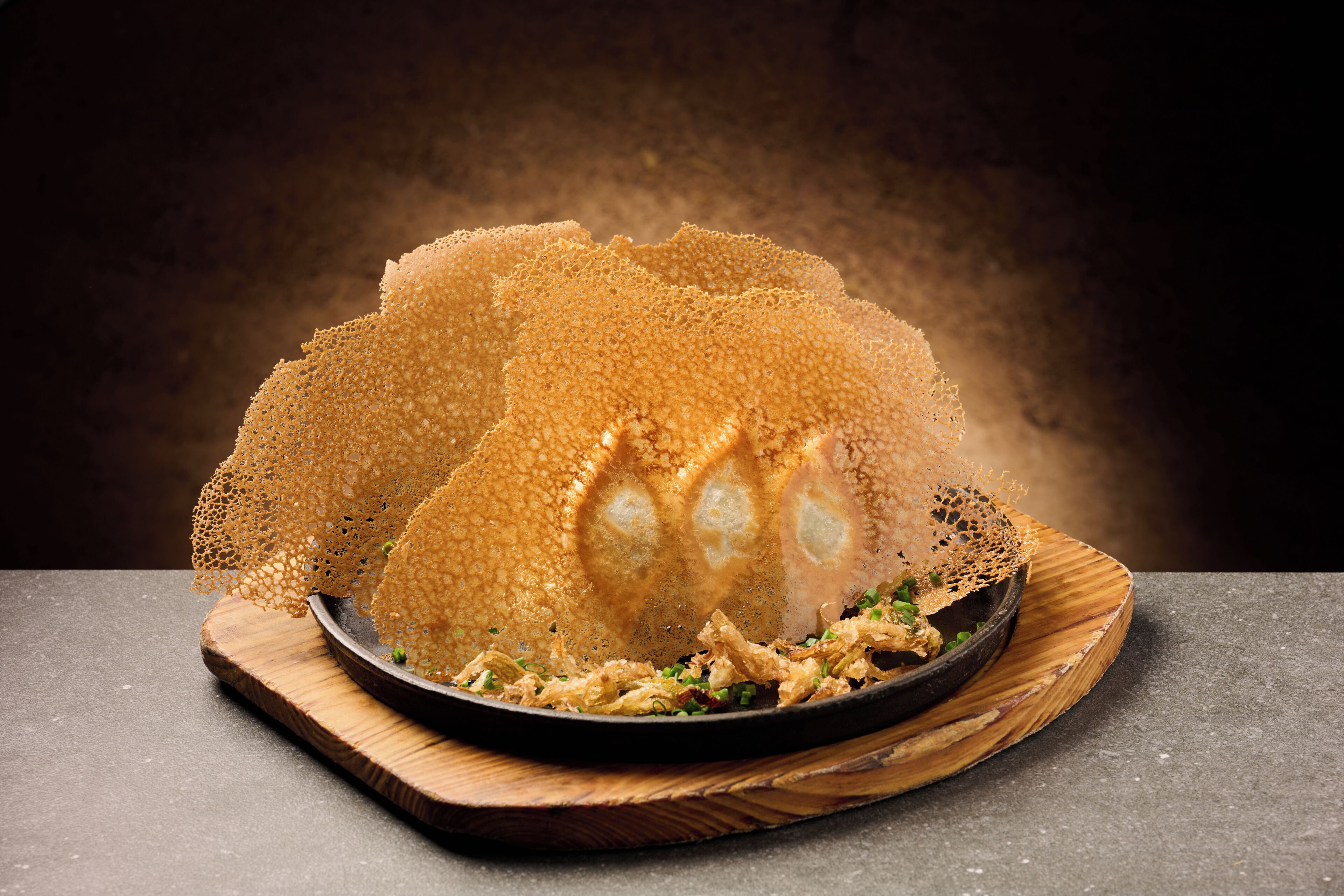 A dish with a crispy, golden-brown texture, possibly a type of fried food, served on a wooden board with a dark background.