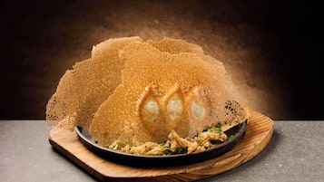 A dish with a crispy, golden-brown texture, possibly a type of fried food, served on a wooden board with a dark background.