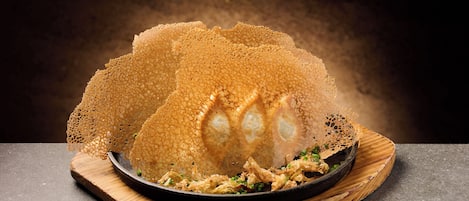 Un plat à la texture croustillante et dorée, probablement un plat frit, servi sur une planche en bois sur fond sombre.