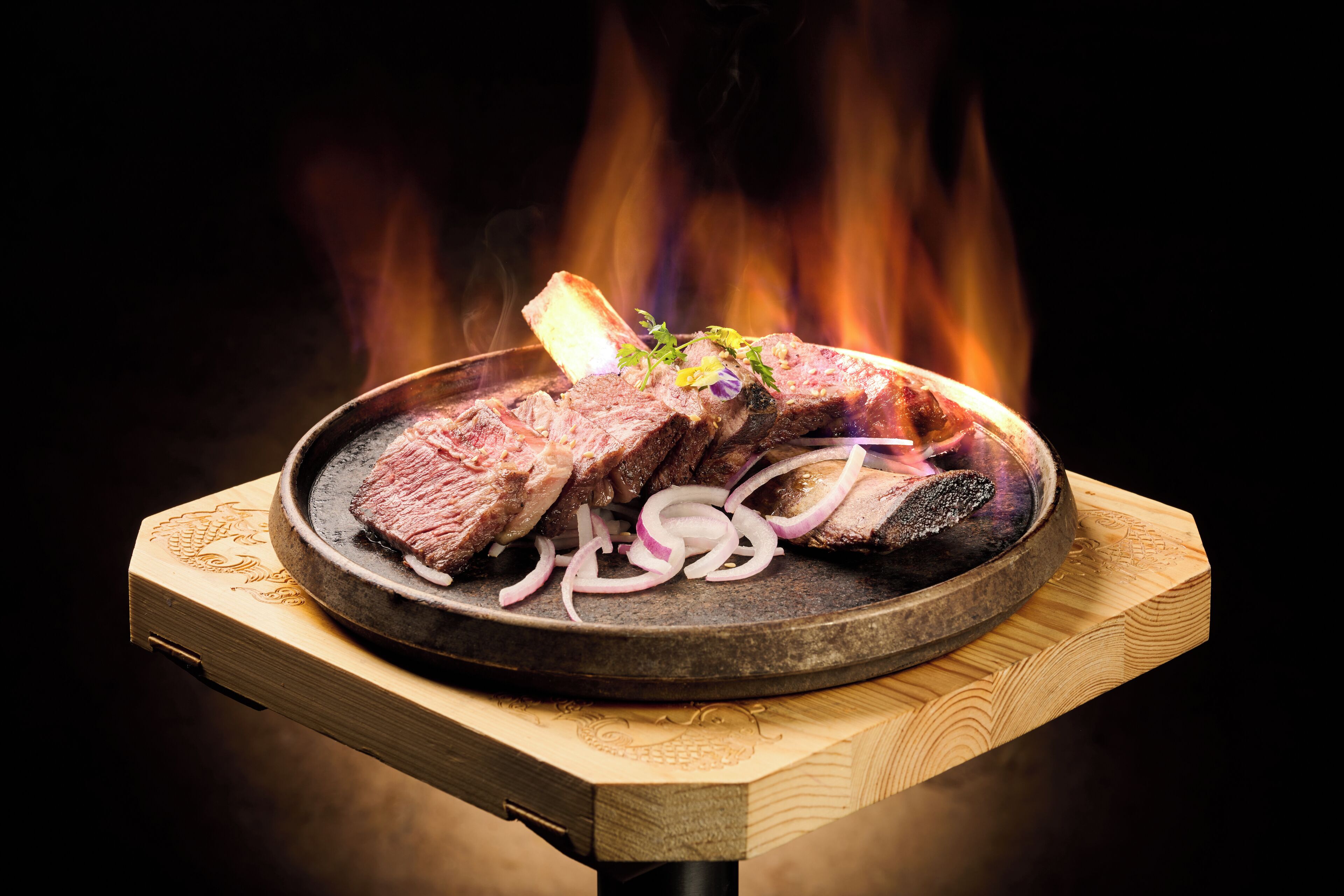 A grilled steak with onions on a wooden board, set against a backdrop of flames.