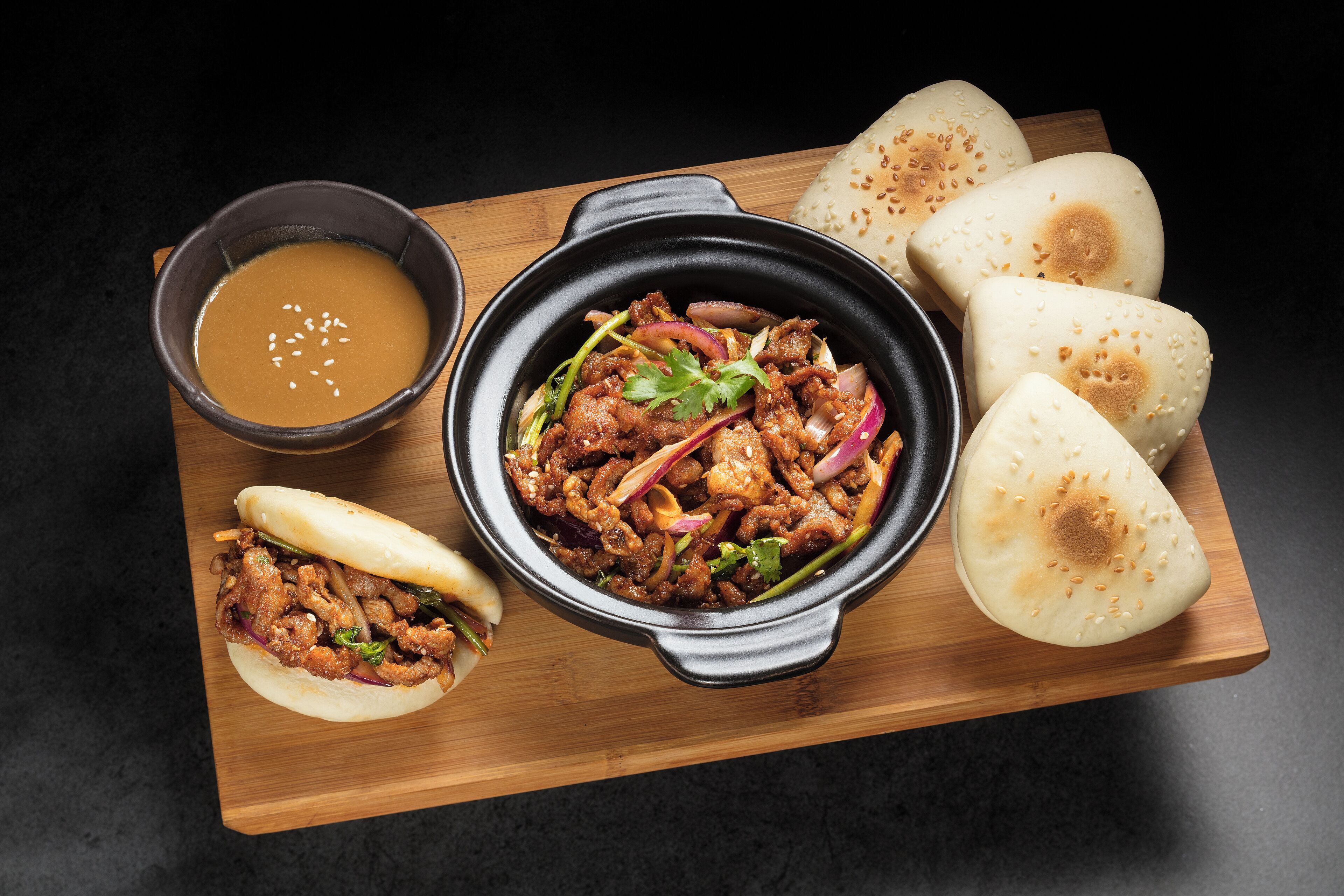 A meal with a bowl of soup, a dish of meat and vegetables, and several pieces of bread on a wooden board.