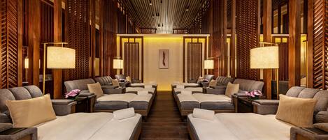 A spa room with multiple massage tables arranged in a symmetrical layout, wooden paneling on the walls, and ambient lighting.
