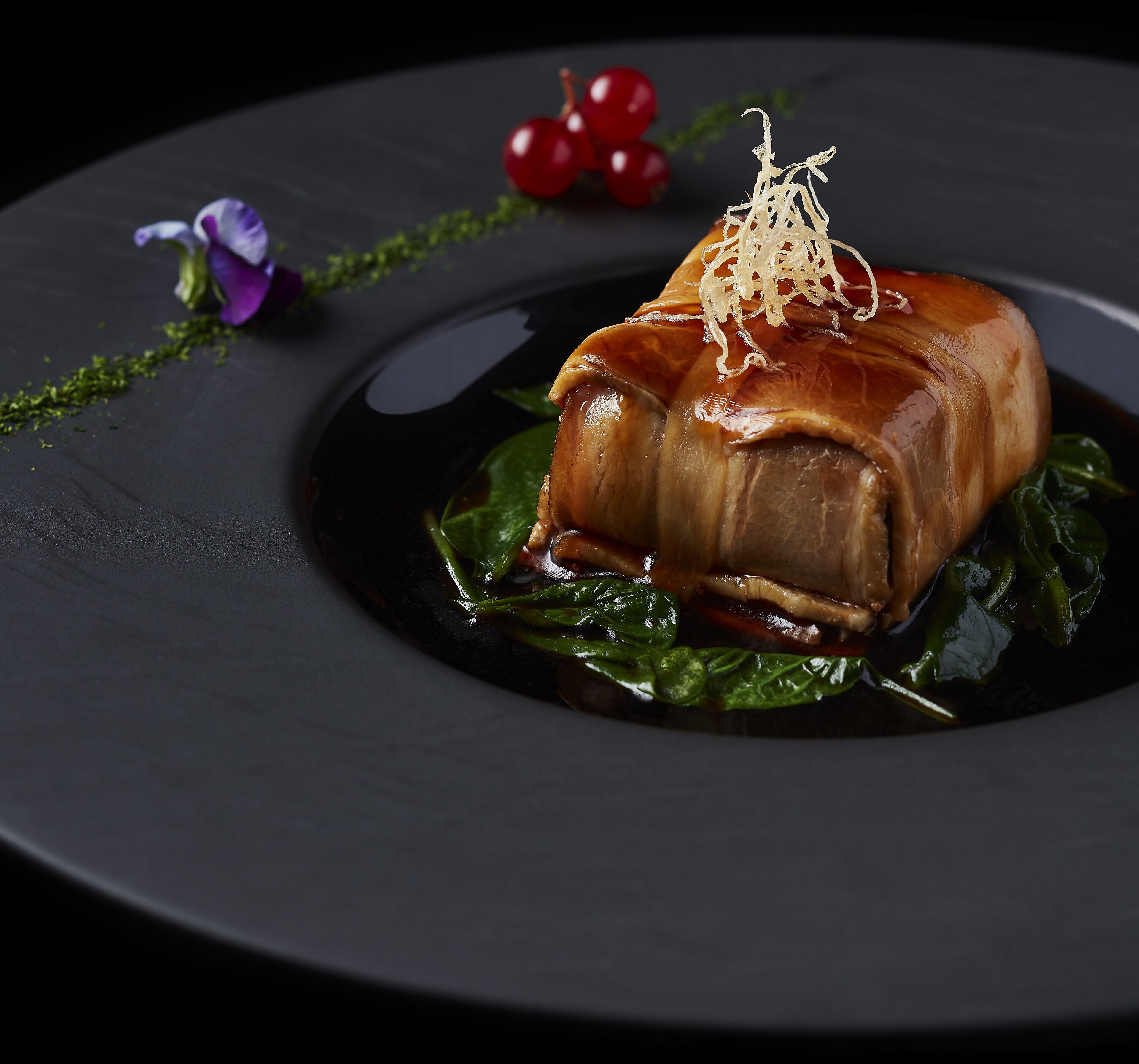 A dish with a glazed meat centerpiece, garnished with shredded ingredients, on a dark plate with greens and red berries.
