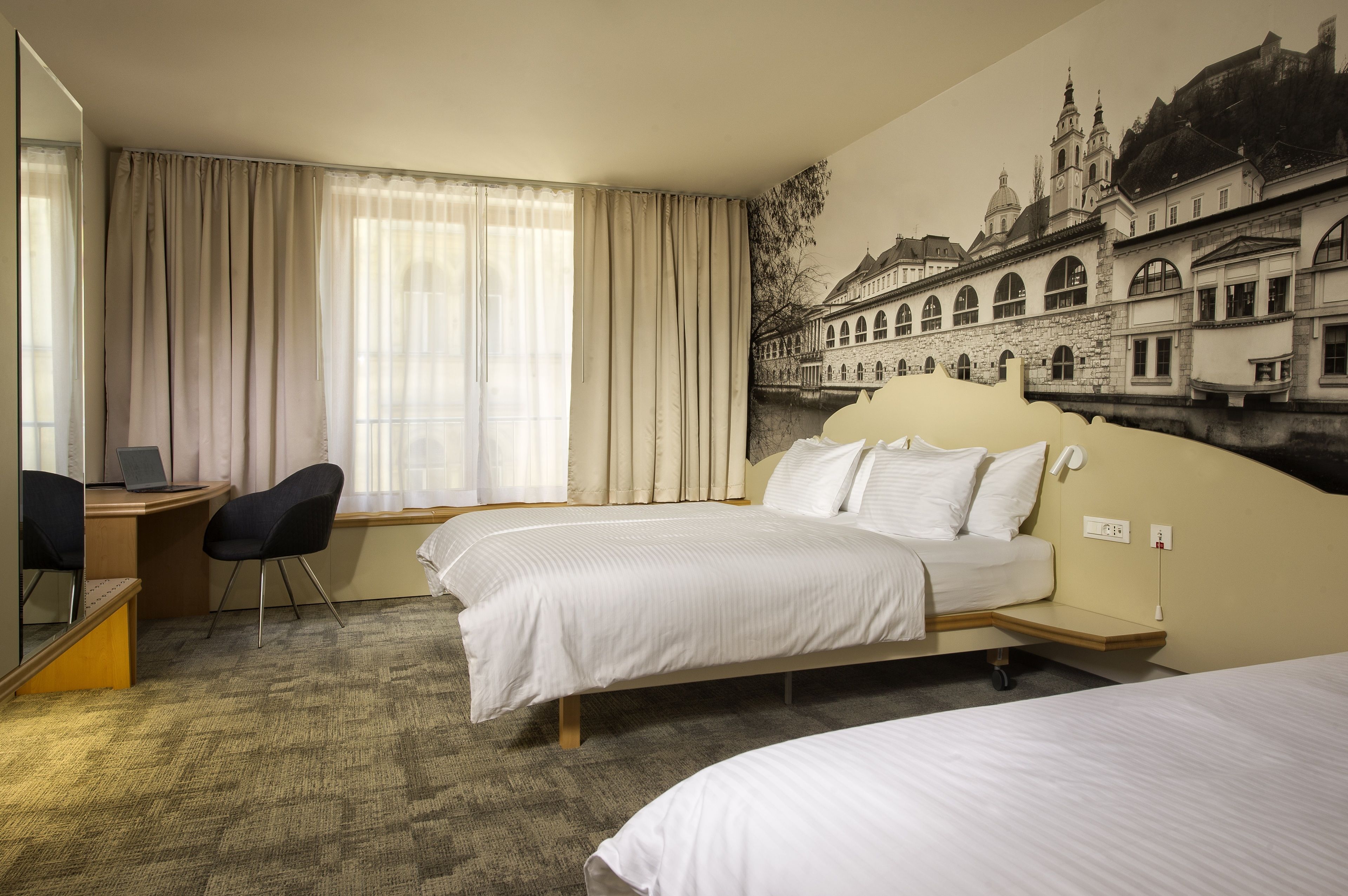 A hotel room with two beds, a desk with a laptop, a chair, and a large window with curtains.