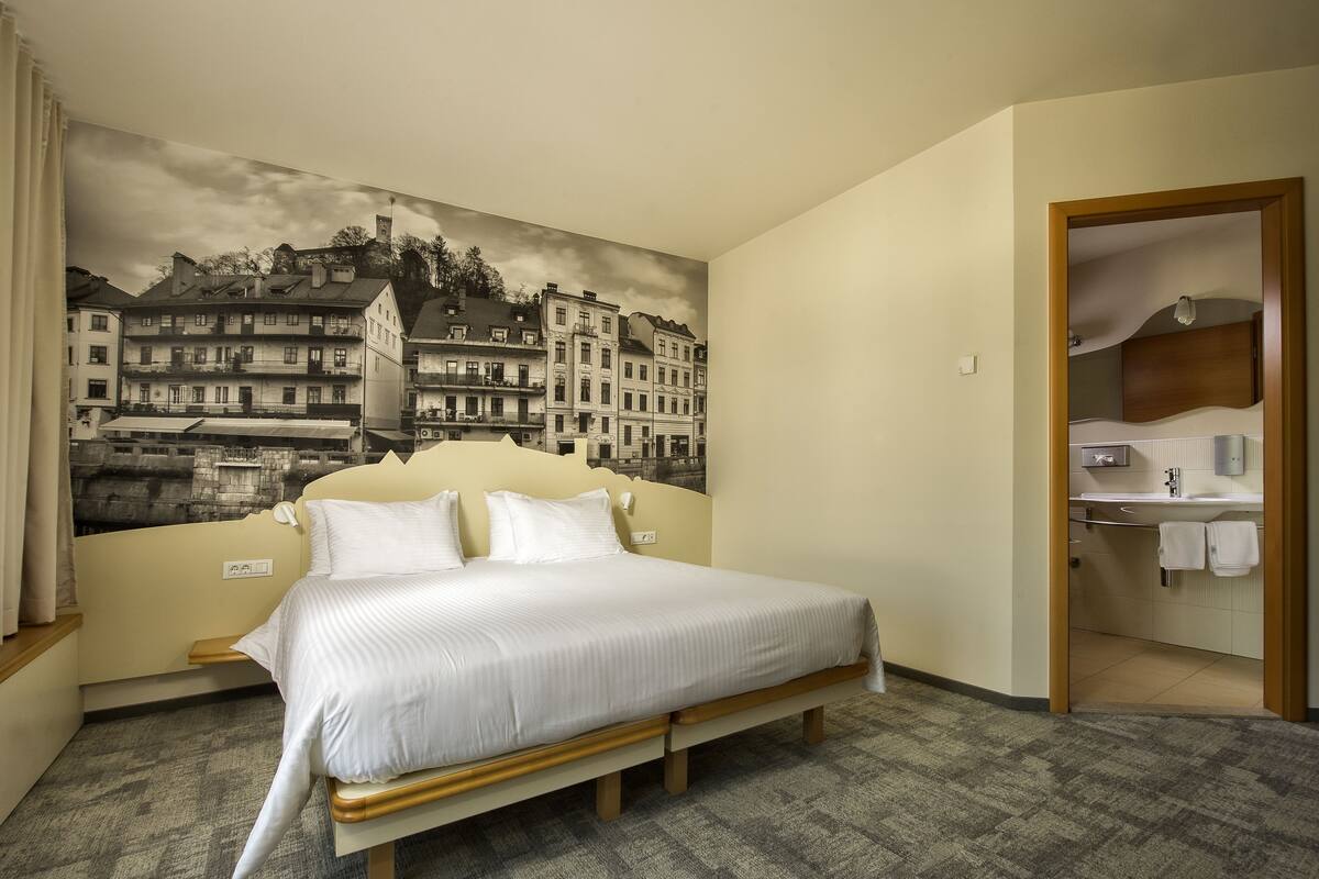 A hotel room with a large bed, a bathroom with a mirror and sink, and a cityscape mural on the wall.