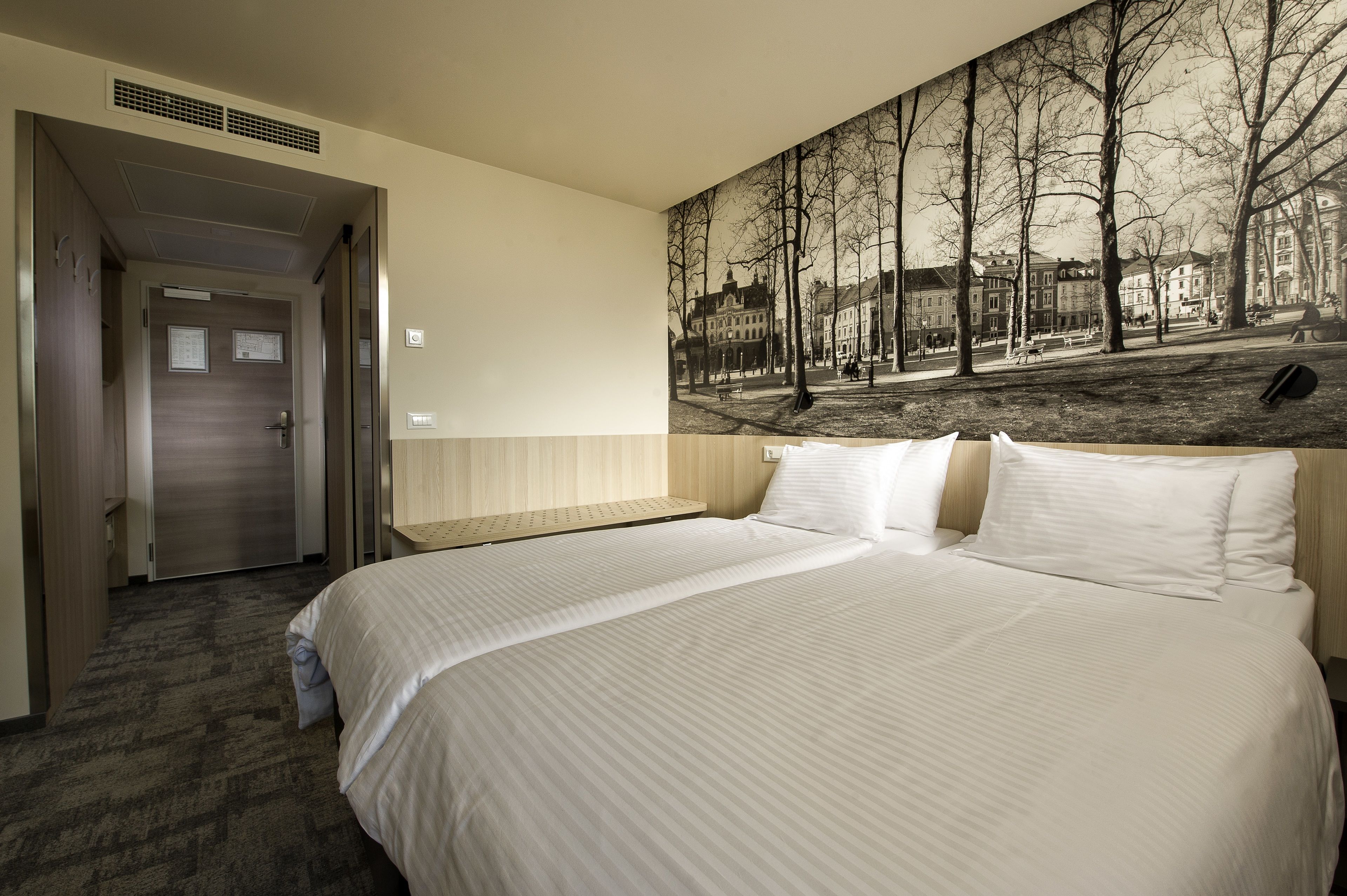 Habitación de hotel con una cama grande, cabecera con un mural de la ciudad, un banco de madera y una puerta que da a un pasillo.