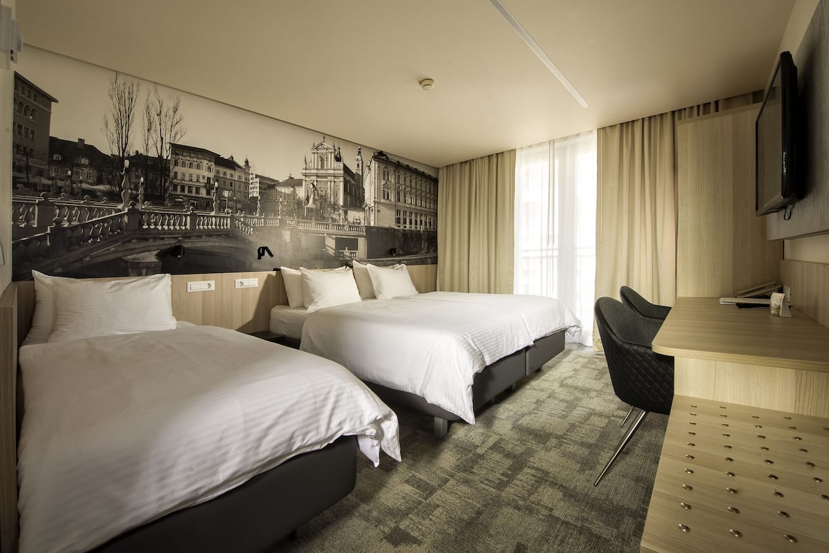 A hotel room with two beds, a desk, a chair, a TV, and a mural of a cityscape.