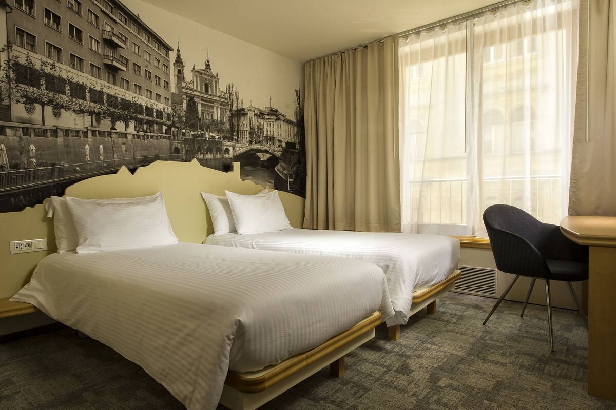 A hotel room with two beds, a large mural of a cityscape, and a window with sheer curtains.
