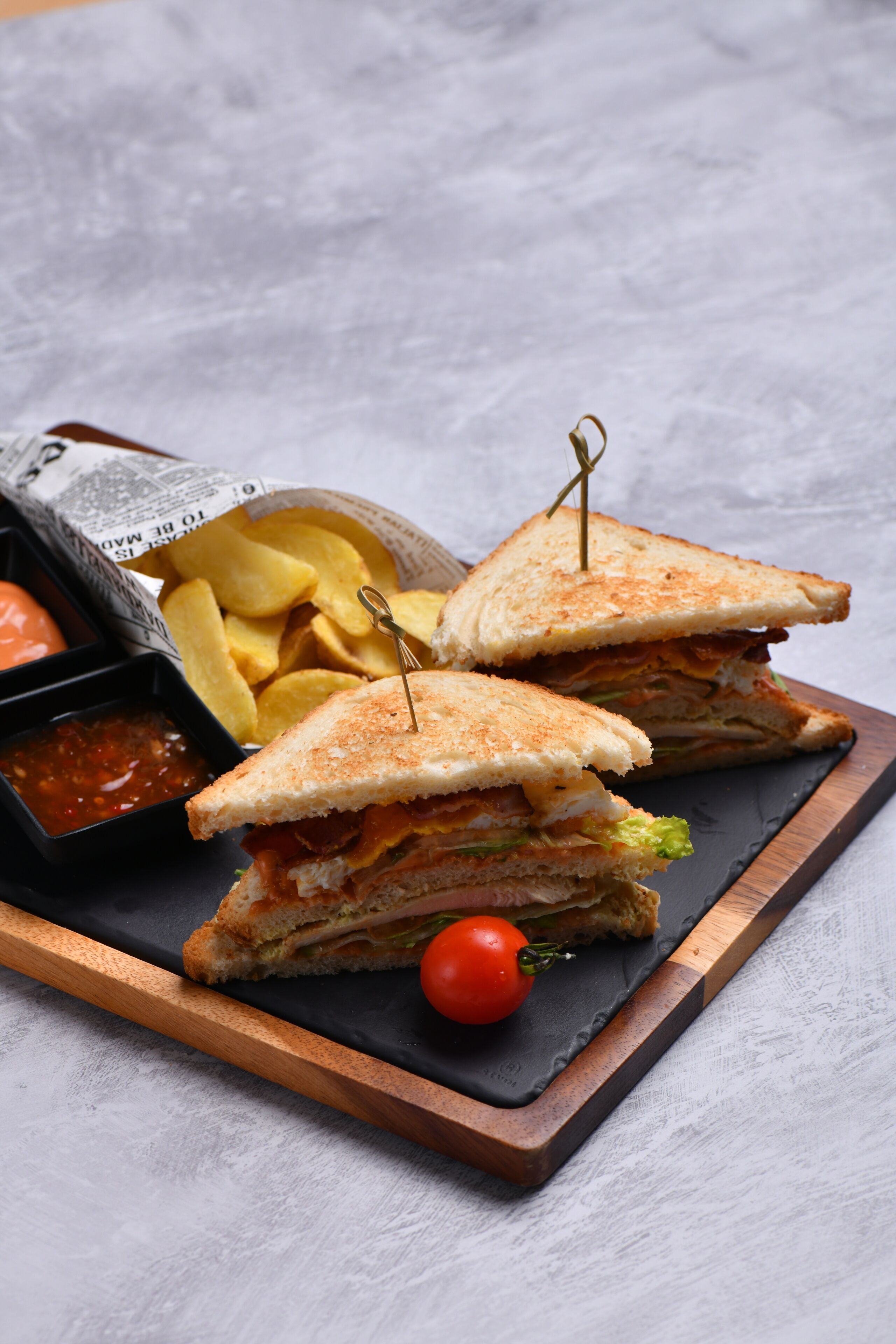 A sandwich cut in half with a toothpick, accompanied by a side of fries and a small bowl of sauce, all placed on a slate board.