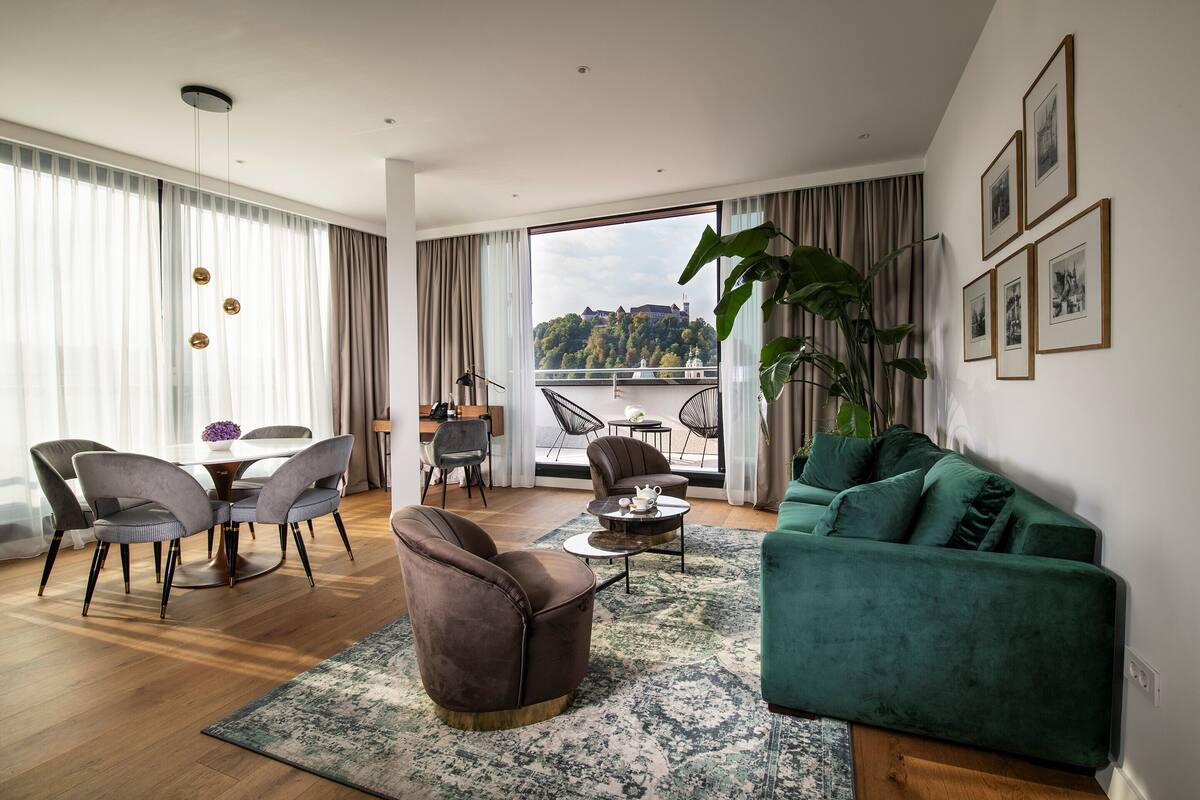 A modern living room with a dining area, large windows with curtains, a green sofa, and a view of a hill.