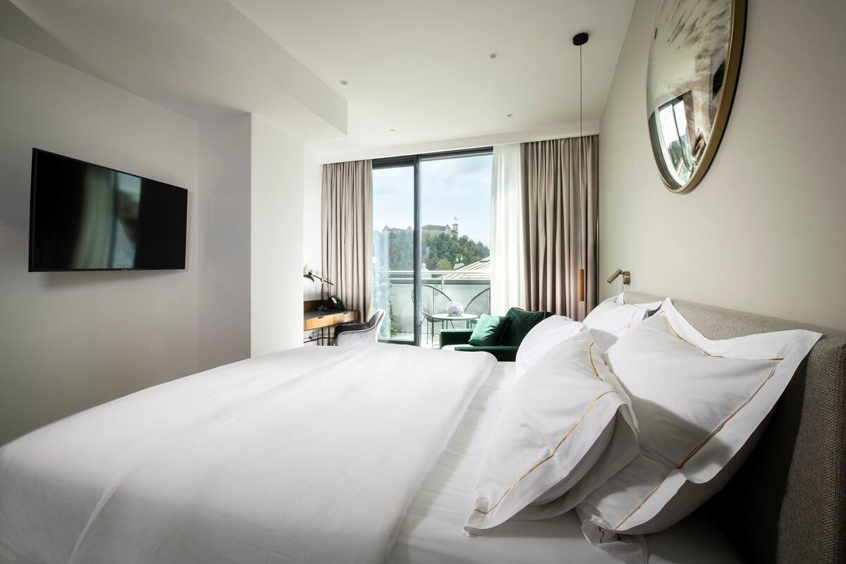 A modern hotel room with a large bed, a flat-screen TV, a desk with a chair, and a balcony with a view of a cityscape.