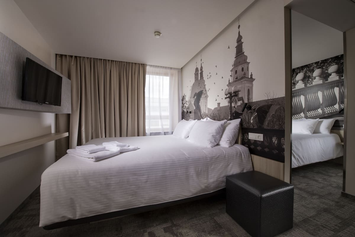 A hotel room with a large bed, a flat-screen TV mounted on the wall, a window with curtains, and a mural of a building.