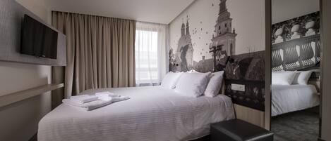 A hotel room with a large bed, a flat-screen TV mounted on the wall, a window with curtains, and a mural of a building.