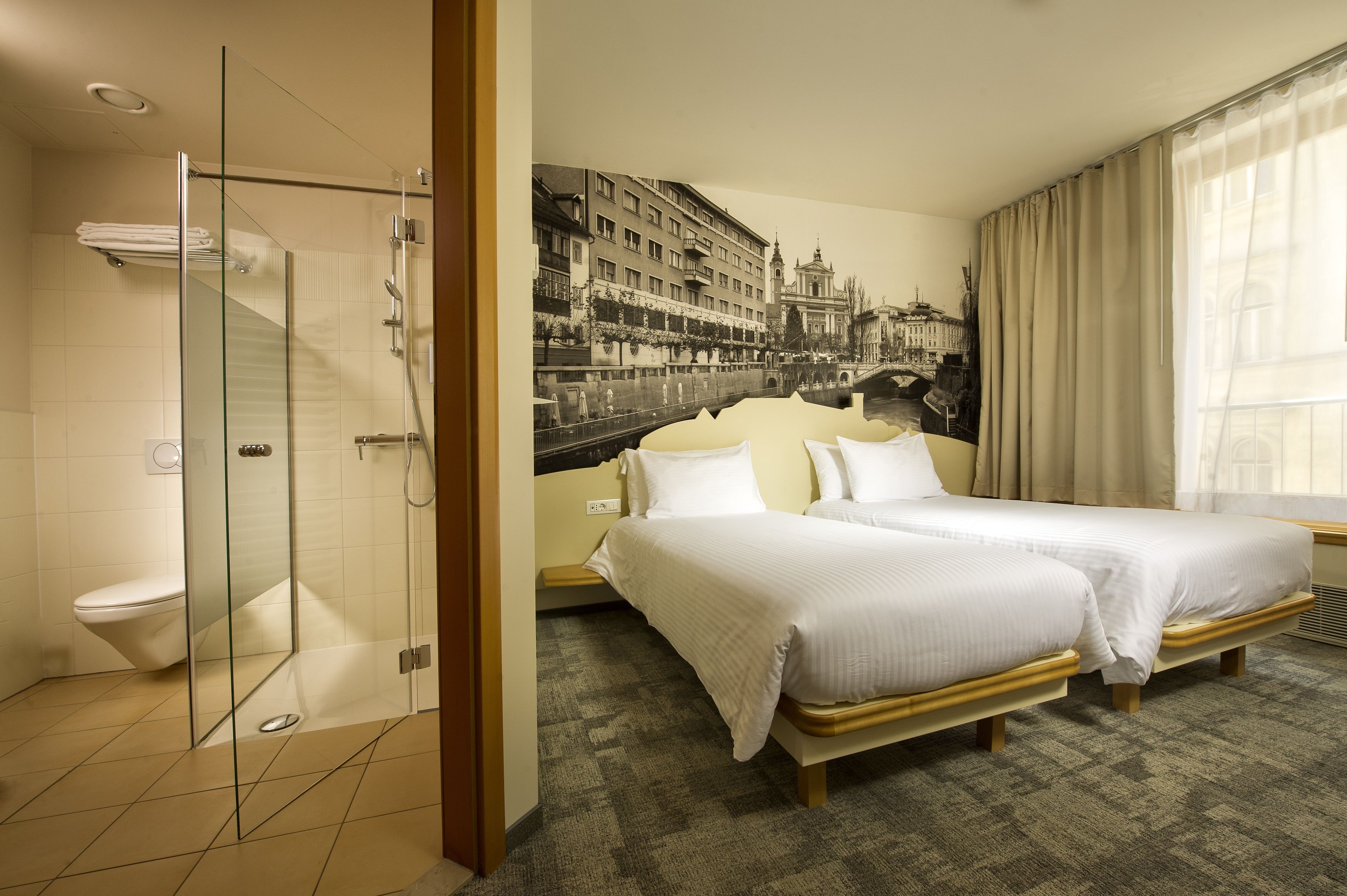 Une chambre d’hôtel dotée d’une douche en verre, d’un lit avec deux oreillers et d’une grande fresque murale représentant un paysage urbain.