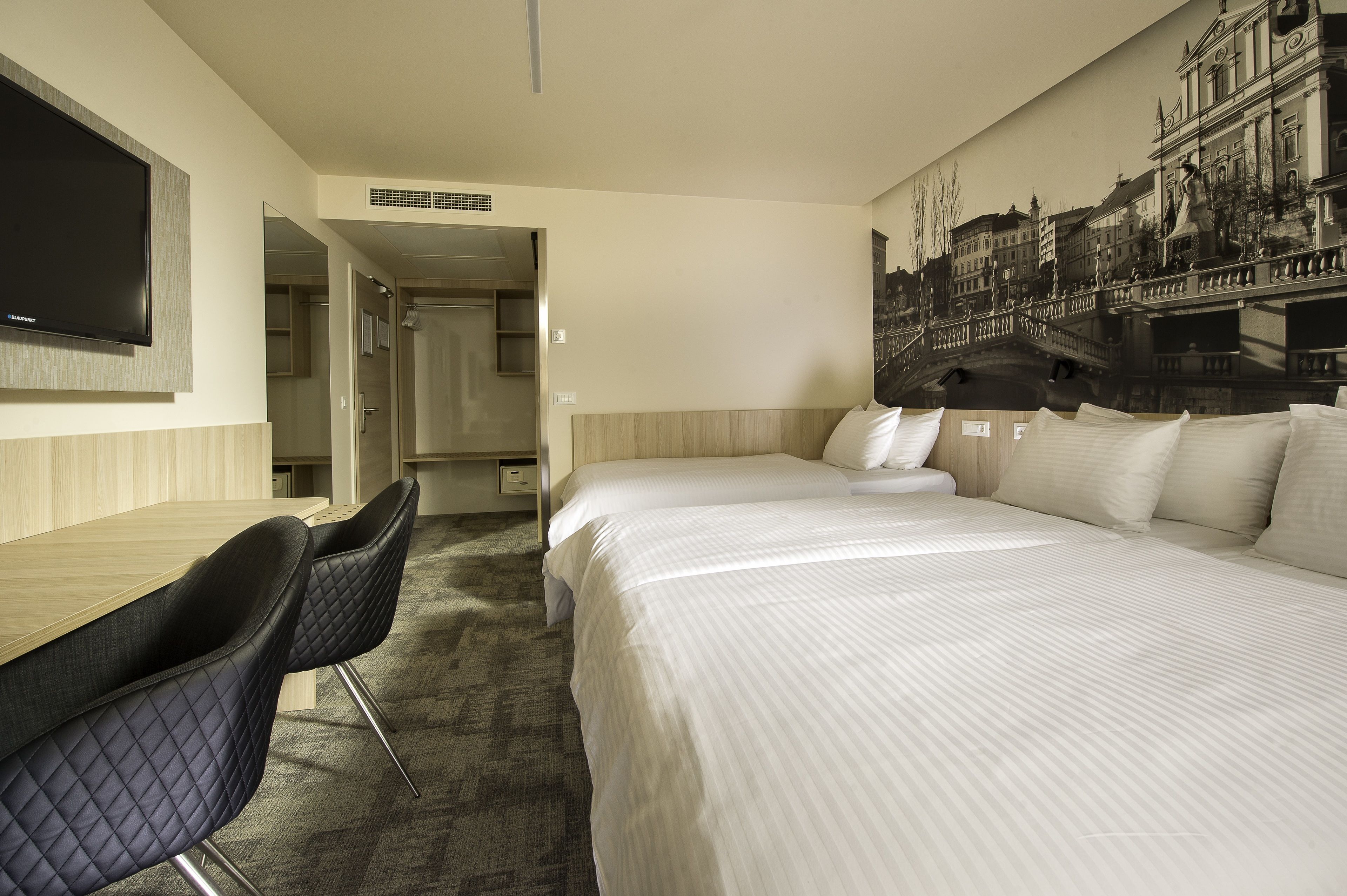 A hotel room with a bed, a desk, and a chair. There is a television mounted on the wall and a painting of a bridge.