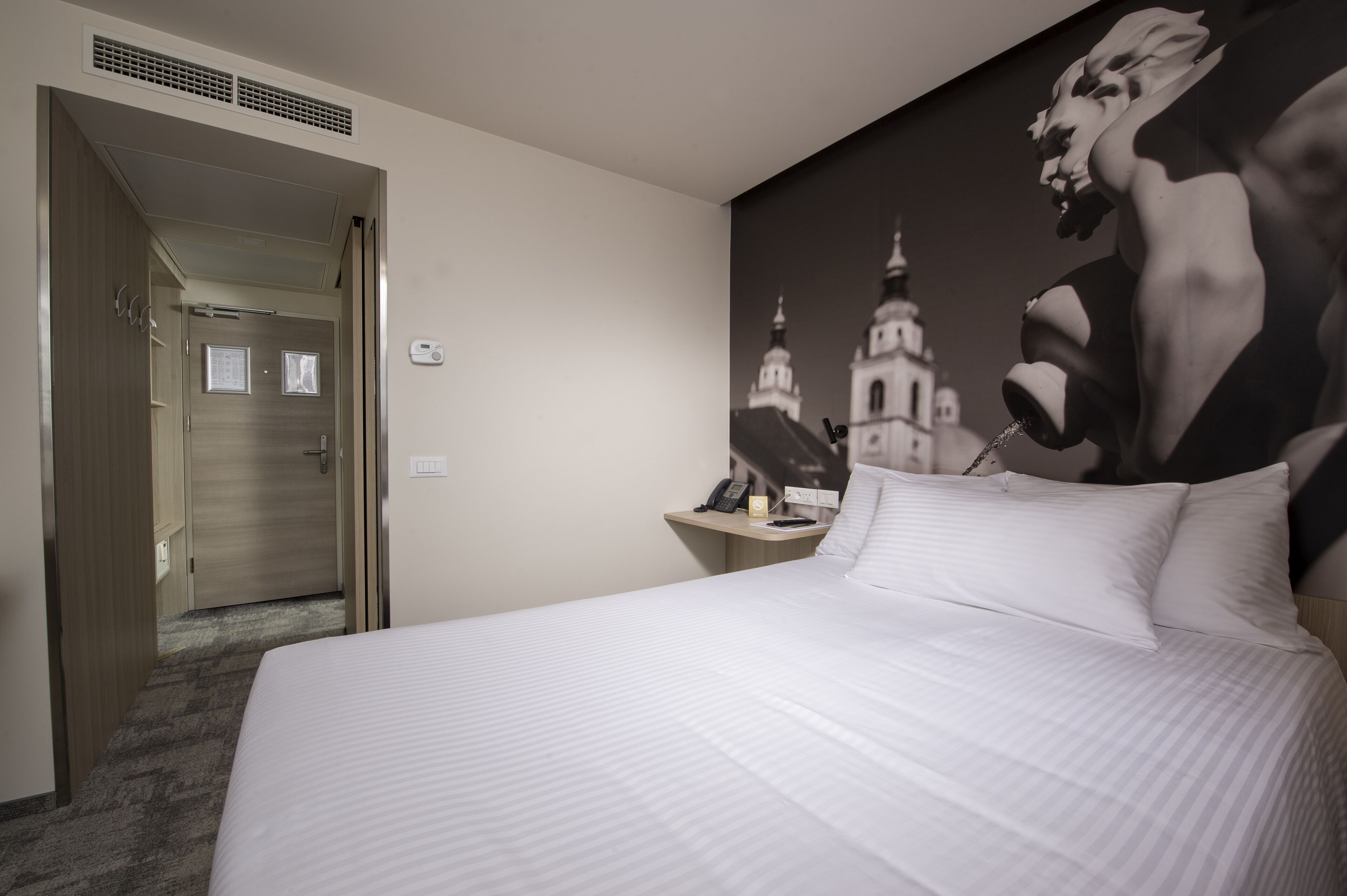 A hotel room with a large bed, a nightstand with a phone and a lamp, a wall-mounted artwork, and a door leading to another room.