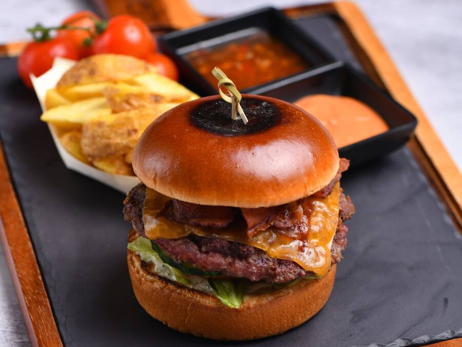Ein Burger mit Zahnstocher, dazu Pommes und eine kleine Soße, alles auf einer Schieferplatte serviert.