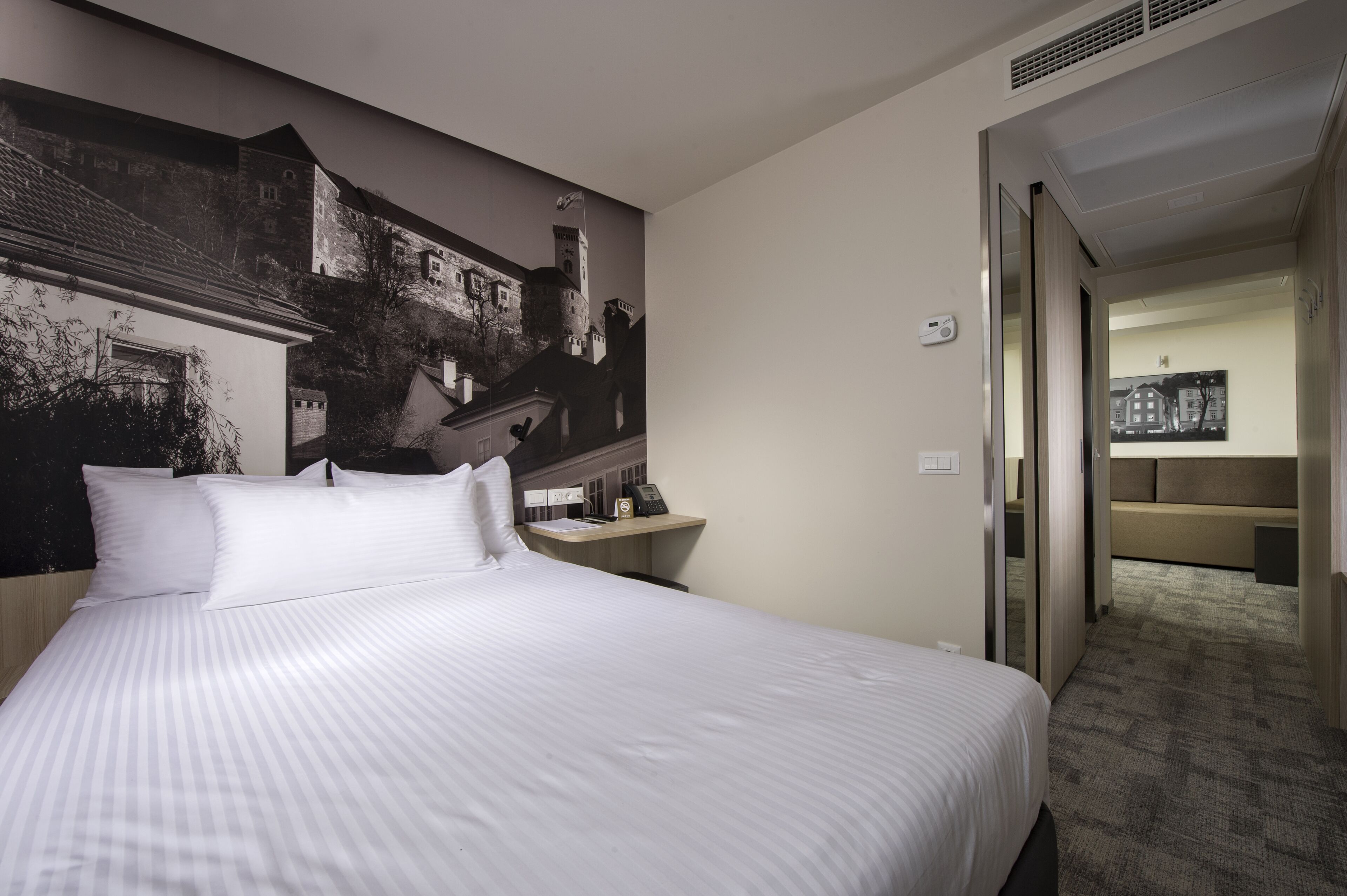 Une chambre d’hôtel avec un grand lit, une table de chevet avec un téléphone et des documents, une œuvre d’art représentant un château accrochée au mur, et un couloir recouvert de moquette menant à une autre pièce.