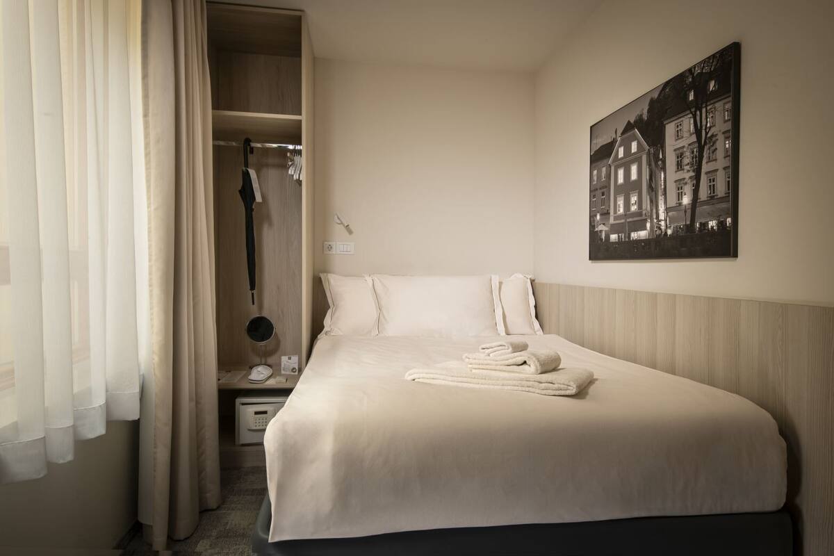 A hotel room with a neatly made bed, a wardrobe, a bedside table with a lamp, and a framed picture on the wall.