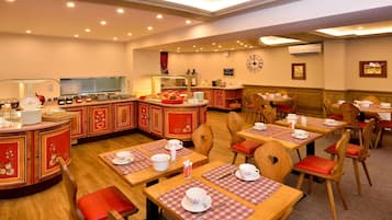 Un restaurante con mesas de madera y sillas rojas con cojines, una zona de cocina y un reloj en la pared.