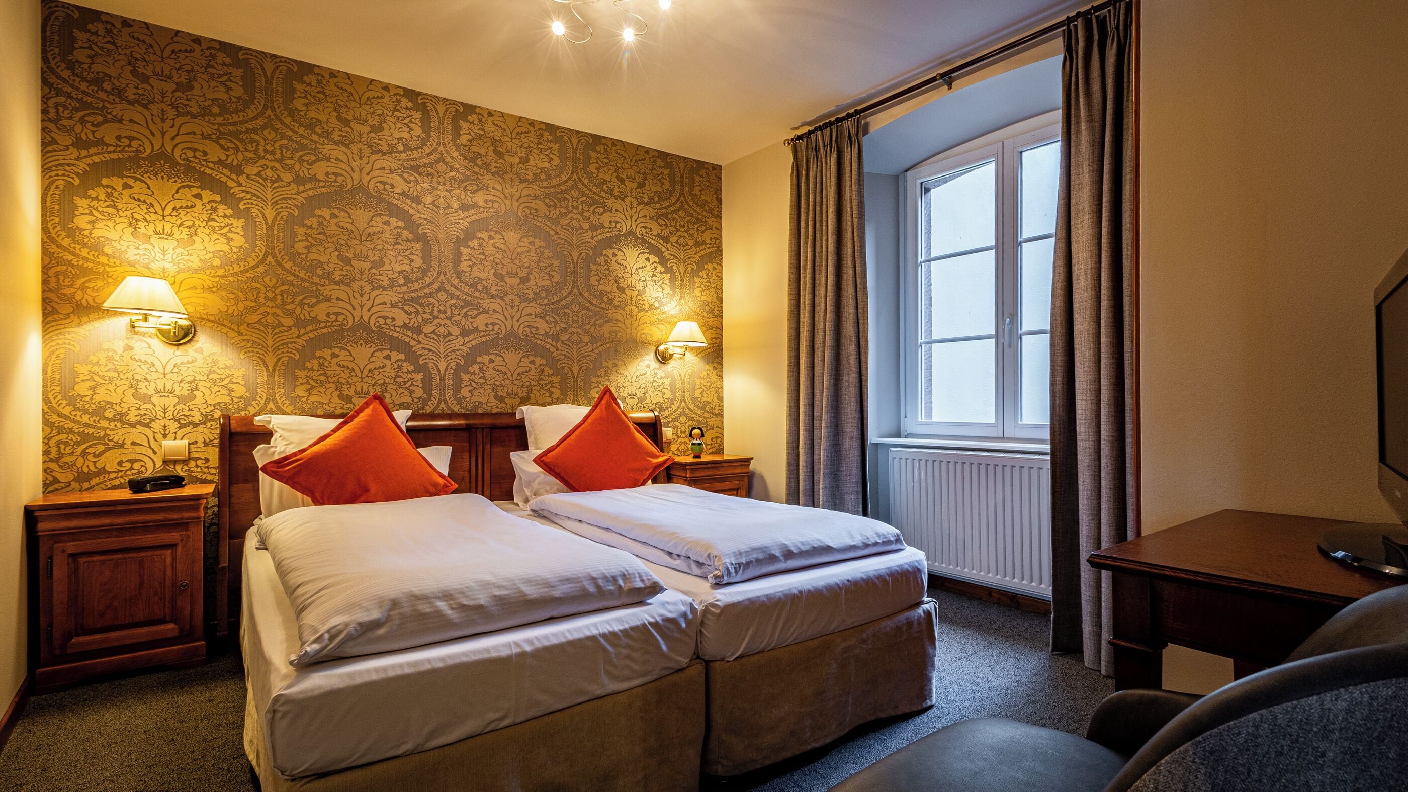 Une chambre d’hôtel avec deux lits, un bureau, une chaise, une fenêtre avec des rideaux et un papier peint à motifs.