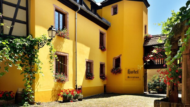 Un bâtiment jaune avec une tour, une enseigne indiquant « hôtel » et des jardinières aux fenêtres.