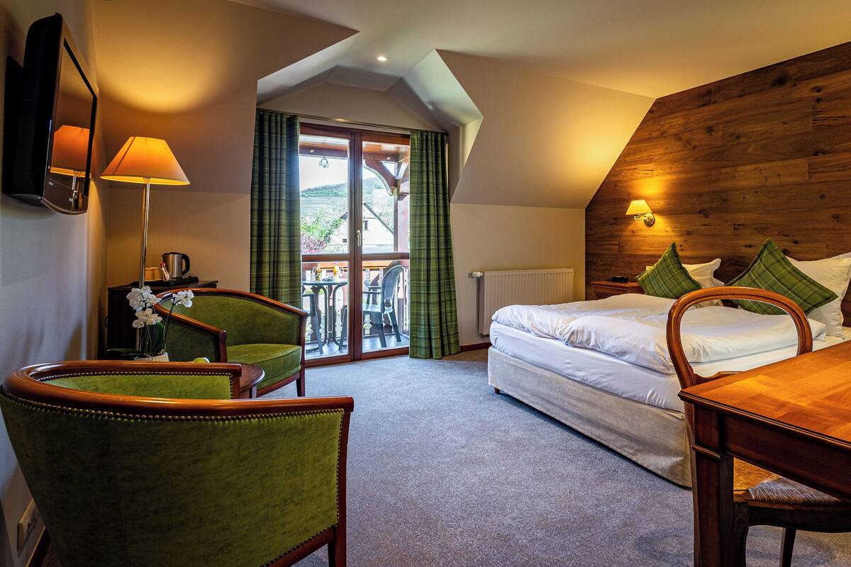 A hotel room with a bed, a desk, a chair, a table, a lamp, a mirror, a window with curtains, and a view of the outdoors.