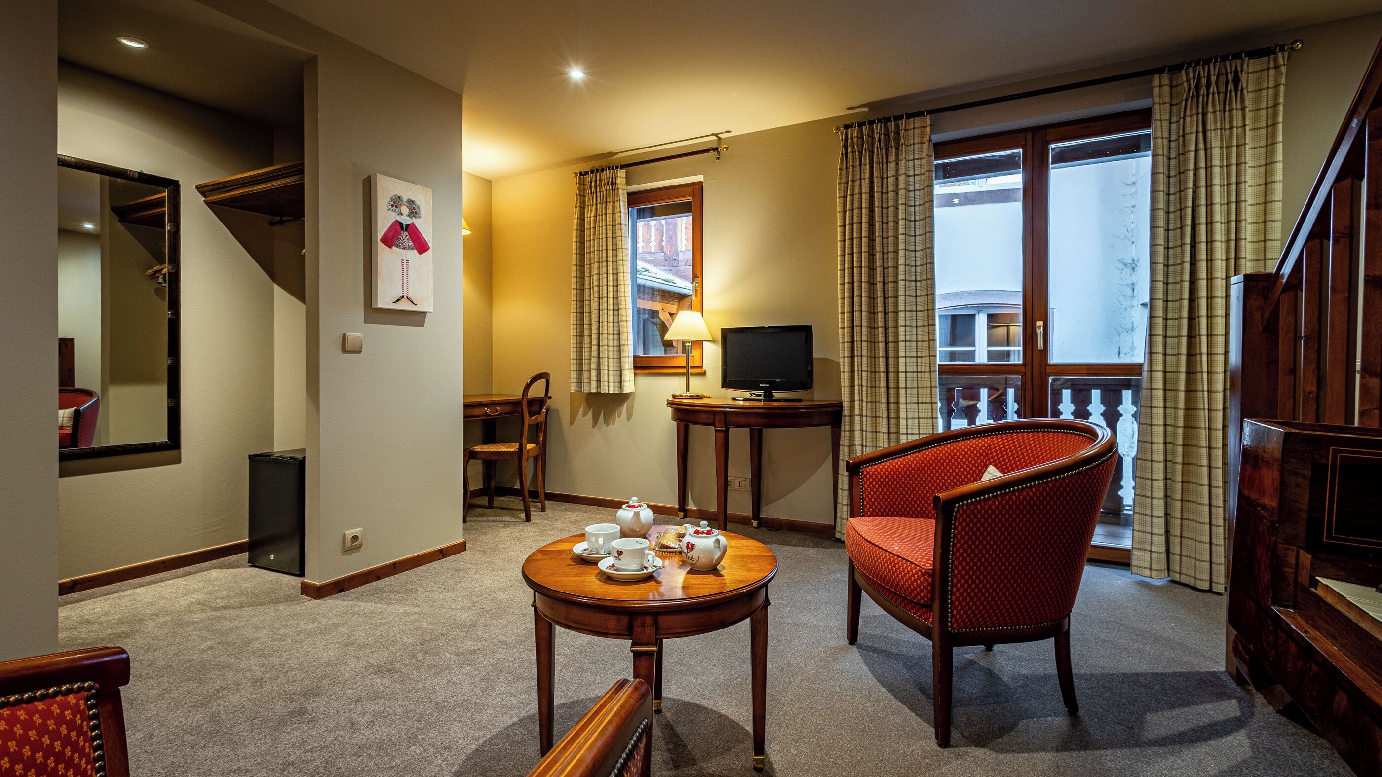 A hotel room with a dining table set for tea, a television, a chair, and a balcony with curtains.