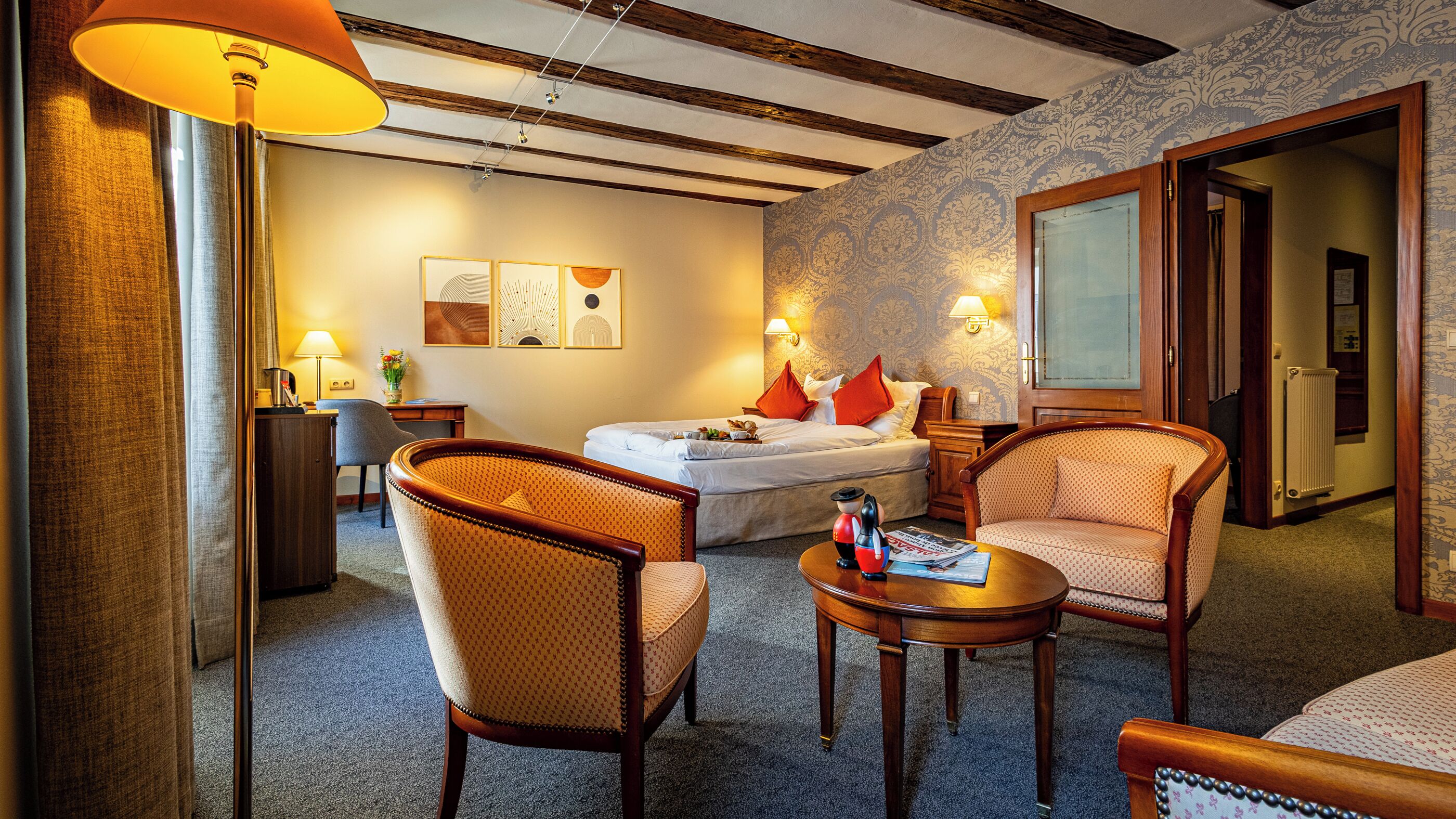 A hotel room with a bed, two armchairs, a small table, a lamp, a desk, and a wall with patterned wallpaper.