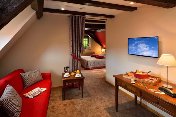 Ein gemütliches Zimmer mit einem roten Sofa, einem kleinen Tisch mit Frühstücksgebäck und -speisen, einem Bett mit roter Bettwäsche und einer Decke mit sichtbaren Balken.