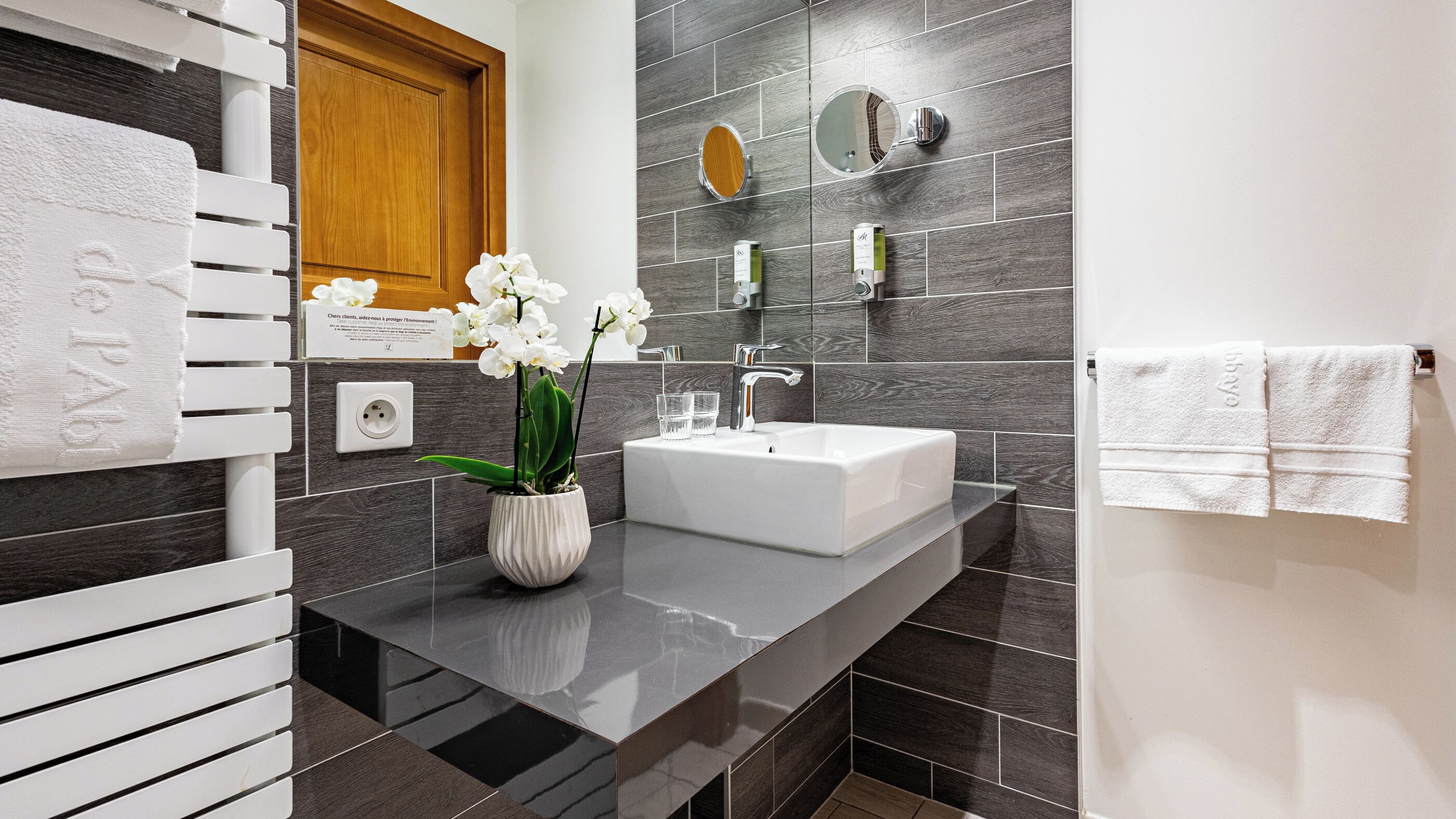 Un baño moderno con un lavabo blanco, baldosas oscuras, una puerta de madera, un toallero blanco y una orquídea en maceta.