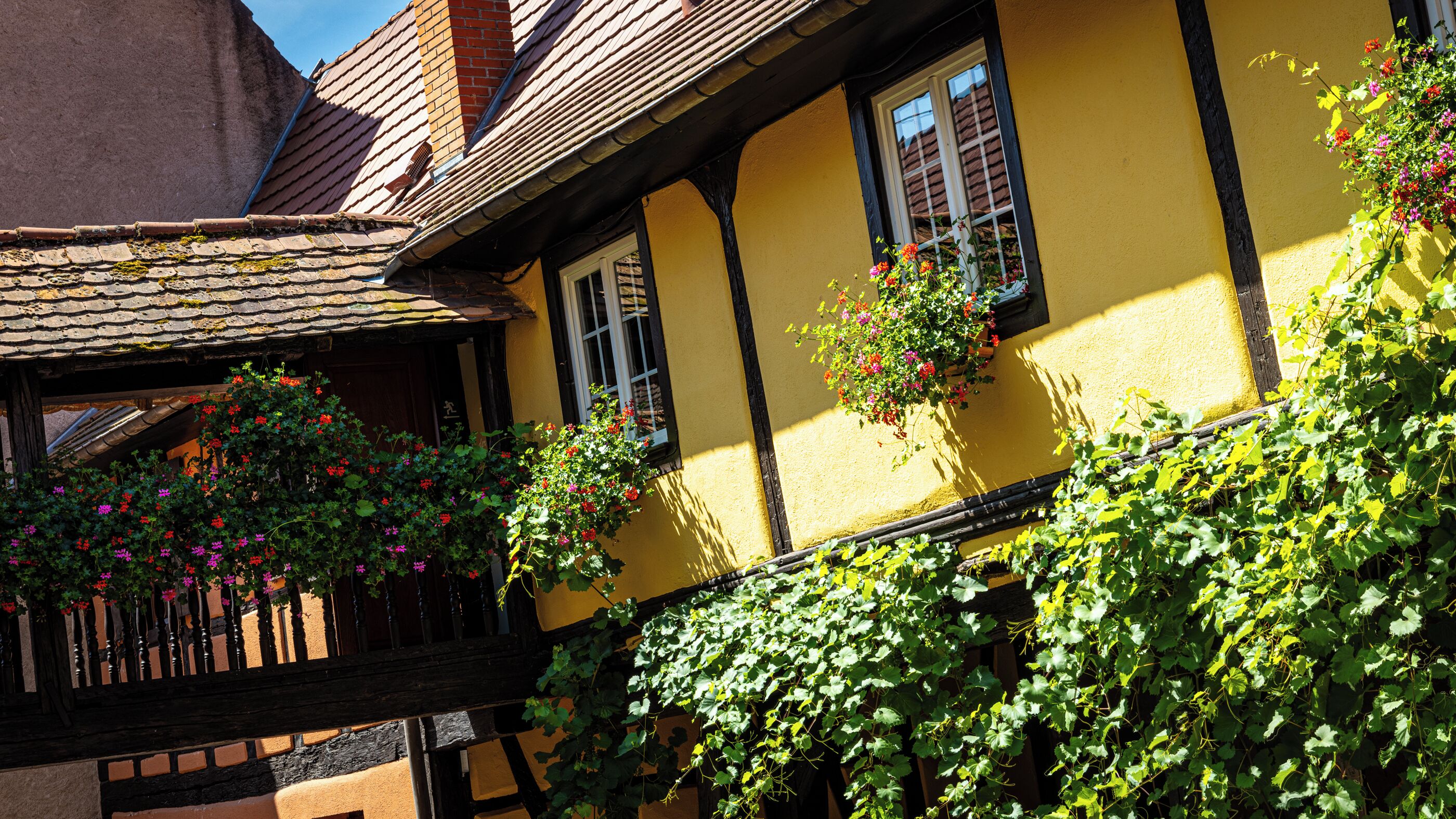 Una casa tradicional con entramado de madera, vigas de madera a la vista, fachada amarilla y macetas con flores en las ventanas.