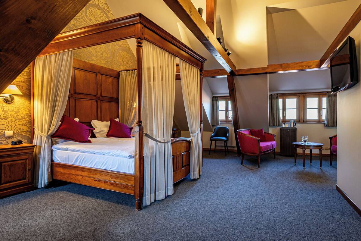 A four-poster canopy bed with a wooden headboard, a small table with glasses, a red chair, and a television mounted on the wall.