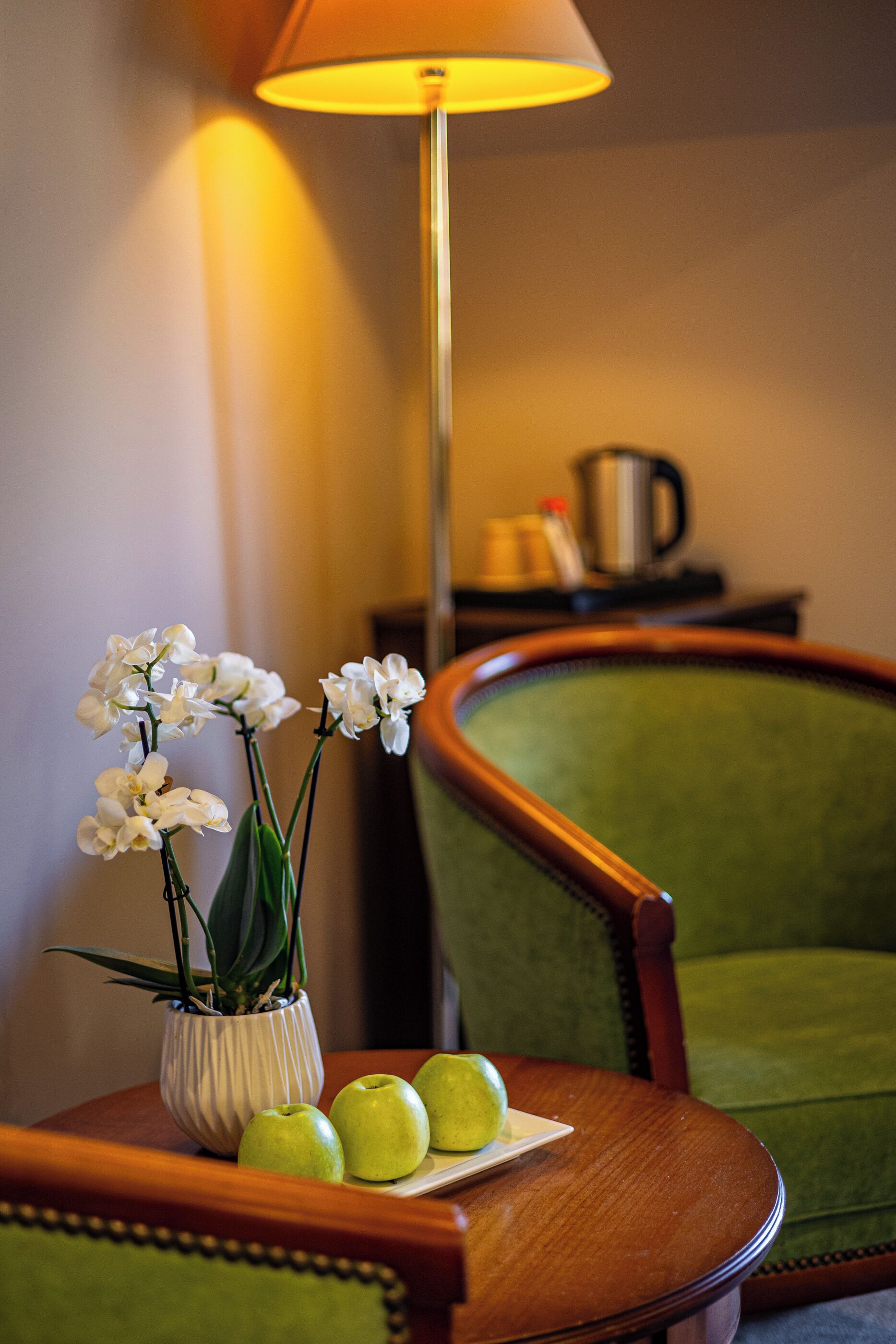 A green upholstered chair, a wooden side table with a vase of white orchids and three green apples, and a floor lamp with a yellow shade.