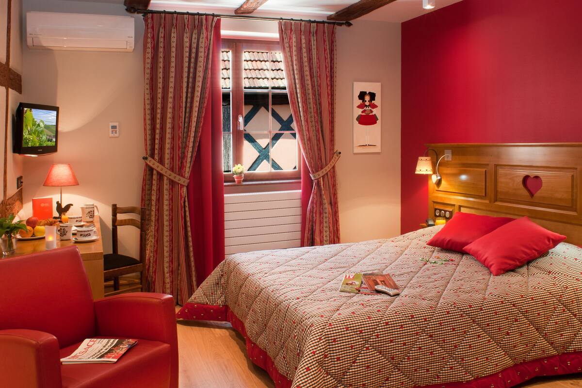 A bedroom with a bed, red walls, a wooden headboard, a window with curtains, a television, and a table with a lamp and a plant.