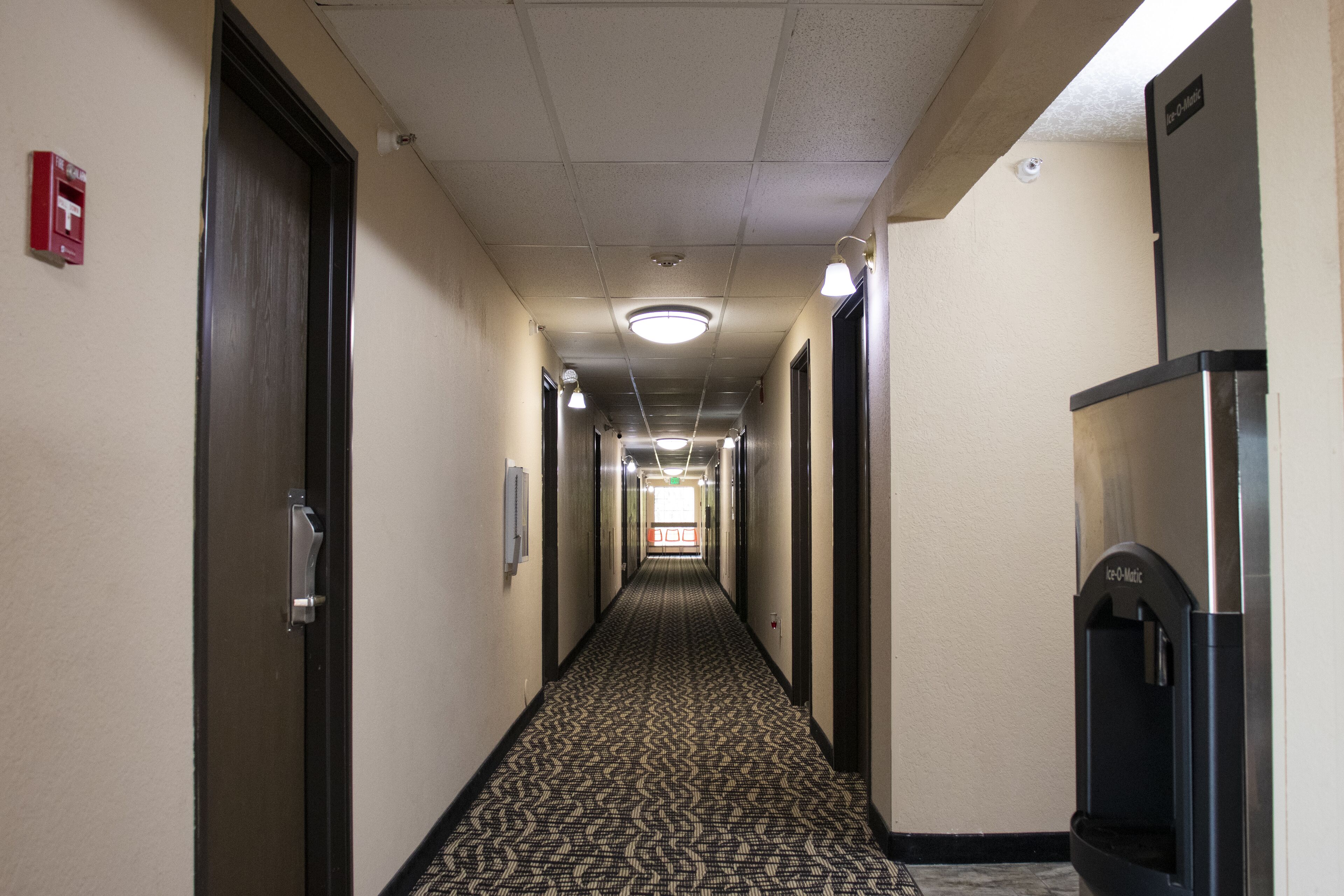 Corredor de hotel com piso carpeteado, portas em ambos os lados e um bebedouro à direita.