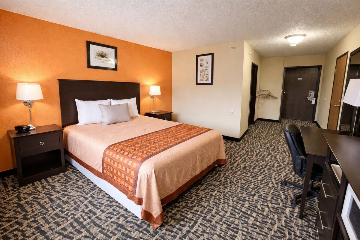 A hotel room with a bed, two bedside tables with lamps, a desk with a chair, and a door.