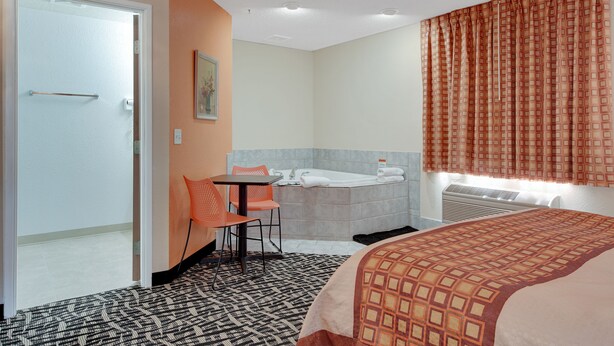 A hotel room with a bed, a bathroom with a bathtub, a table with two chairs, and a painting on the wall. - North Town Inn & Suites (Finlayson)
