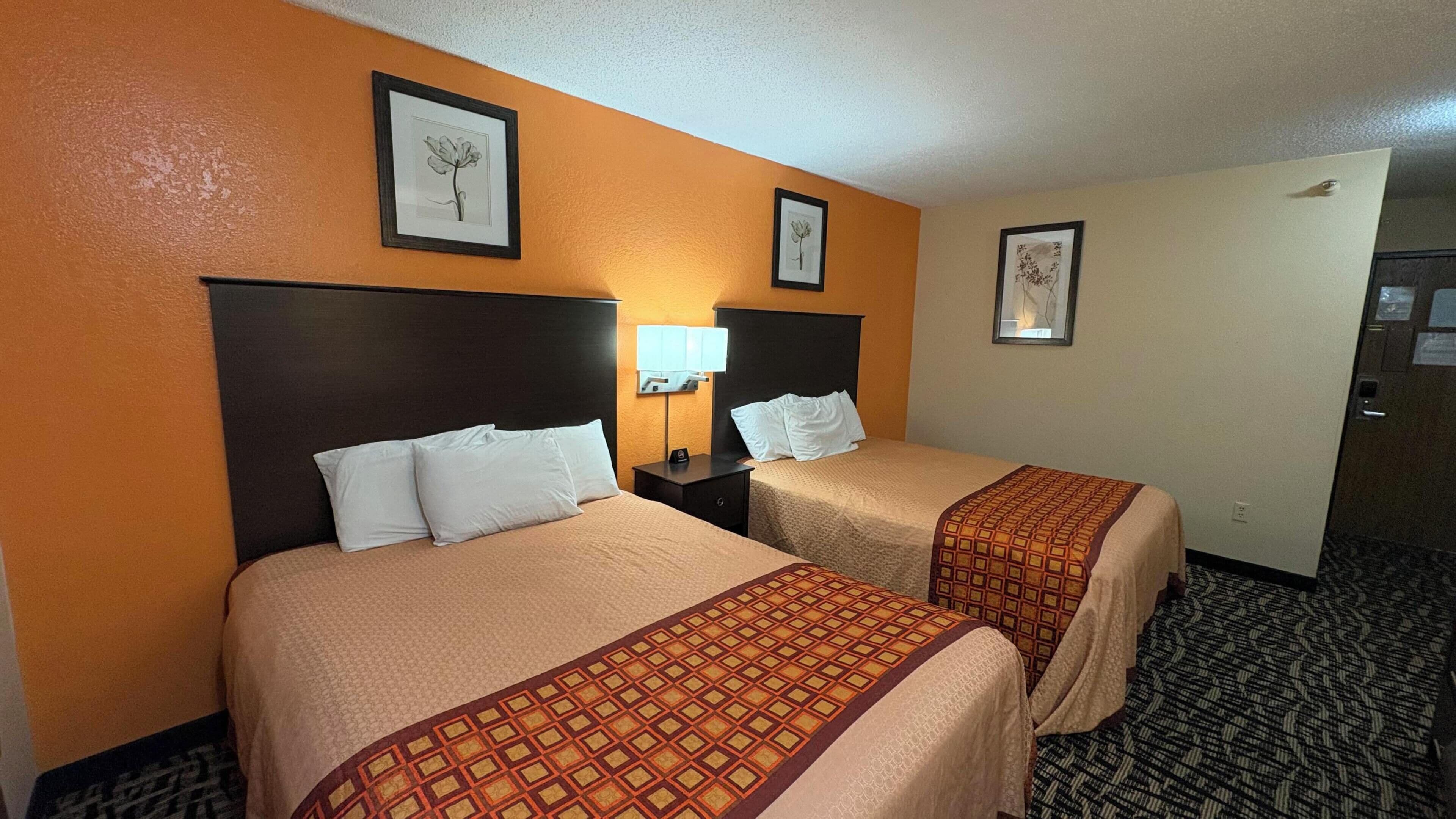 A hotel room with two beds, a nightstand with a lamp, and framed artwork on the walls.