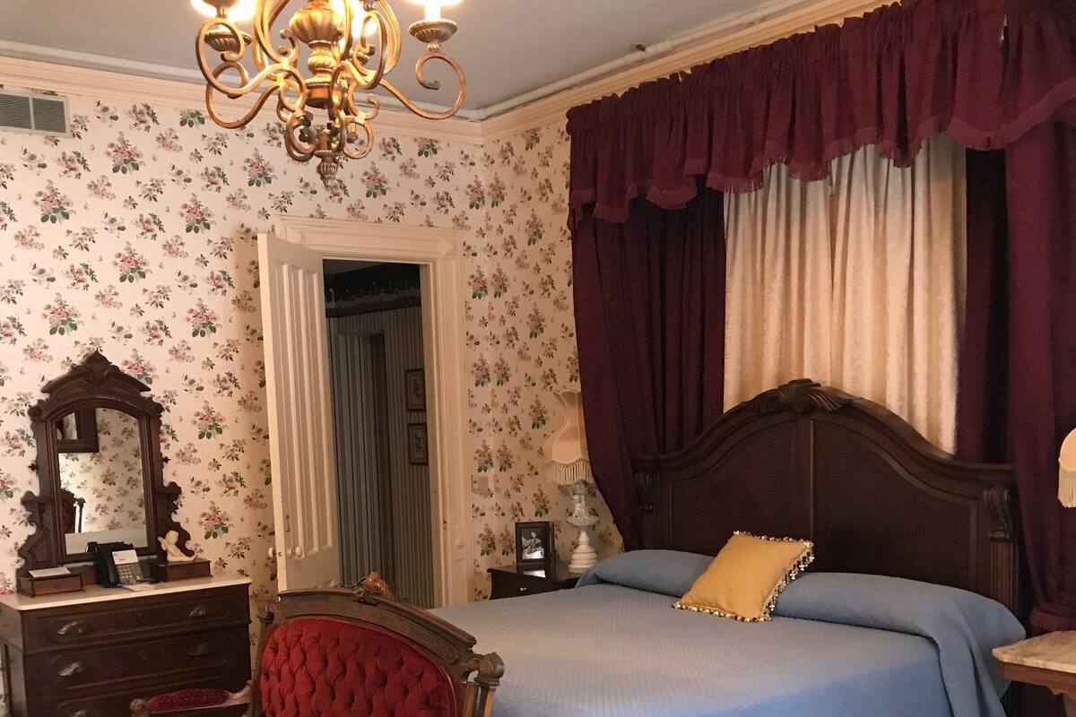 A bedroom with a large bed, a red upholstered chair, a wooden nightstand, a dresser, a mirror, and a chandelier.