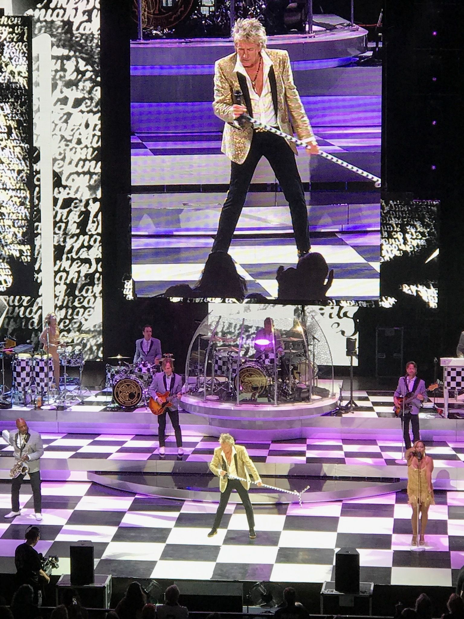A stage with a checkered floor, a band performing, and a large screen displaying a performer.